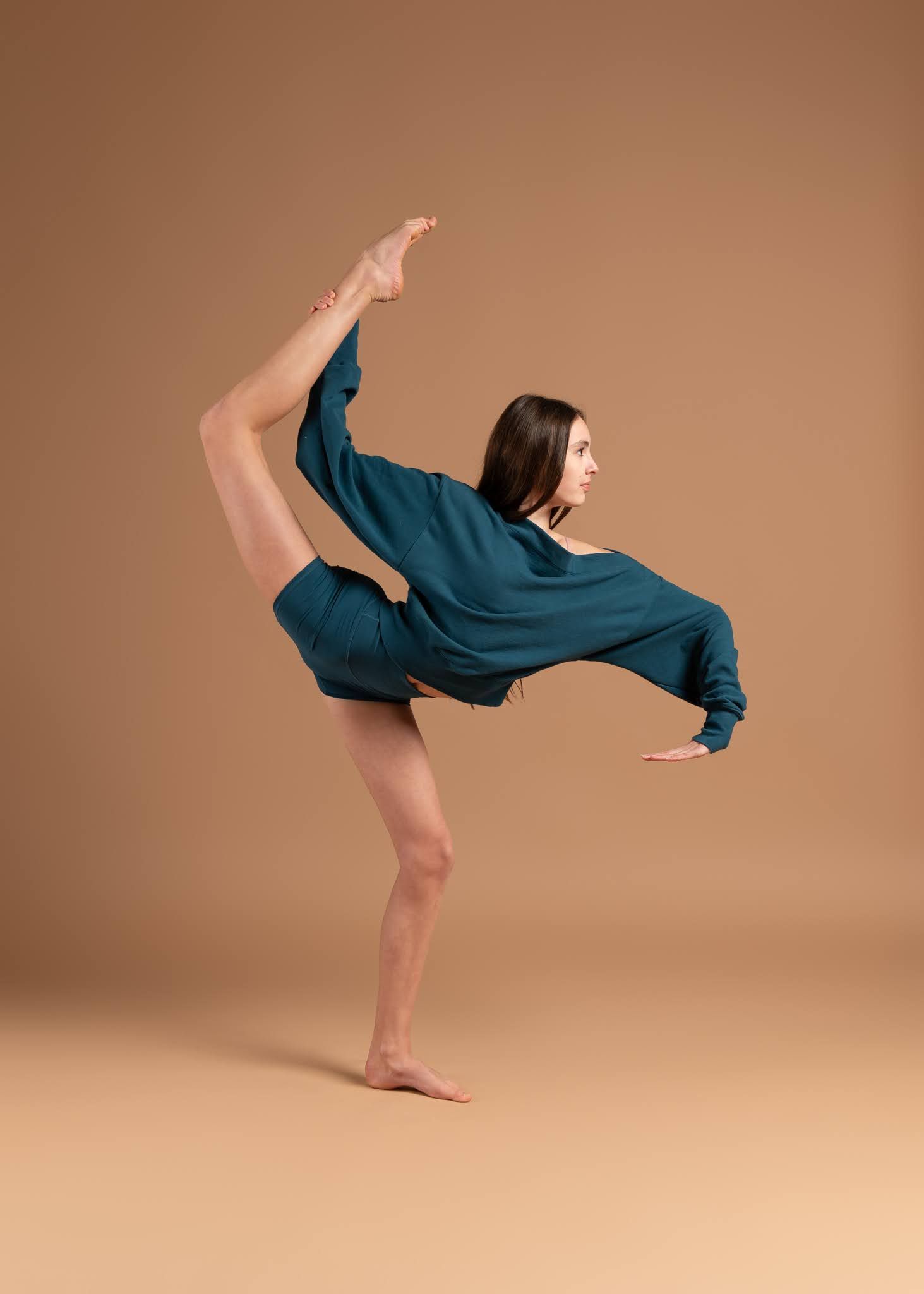 A female dancer performs a stunning standing split in a teal outfit against a warm beige background.