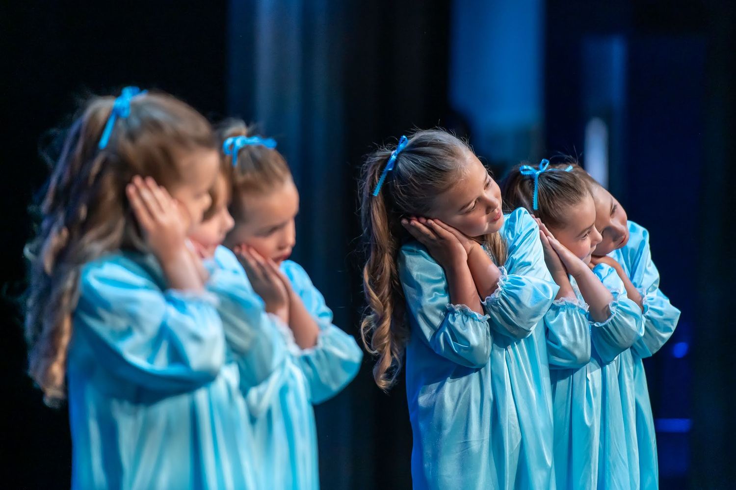 Four young girls in matching light blue dresses perform a synchronized dance routine on stage.