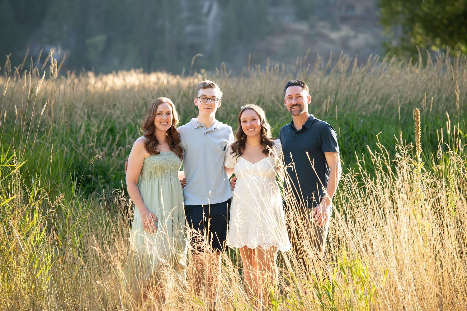Family of four smiling together in tall golden grass field in bright summer sunlight.