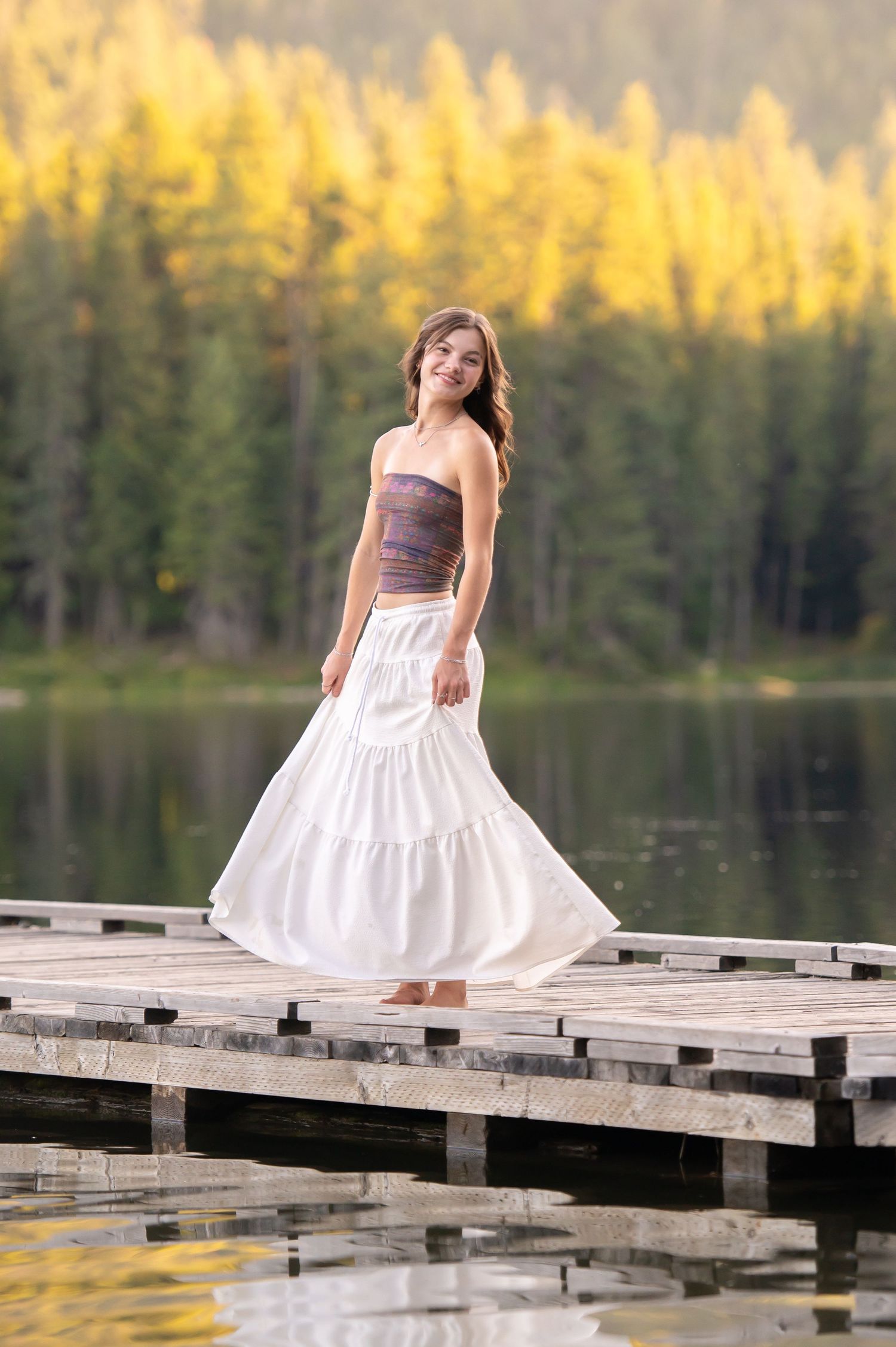Young woman in white tiered skirt and purple strapless top twirling on a wooden dock by a forest lake in the Idaho Panhandle.