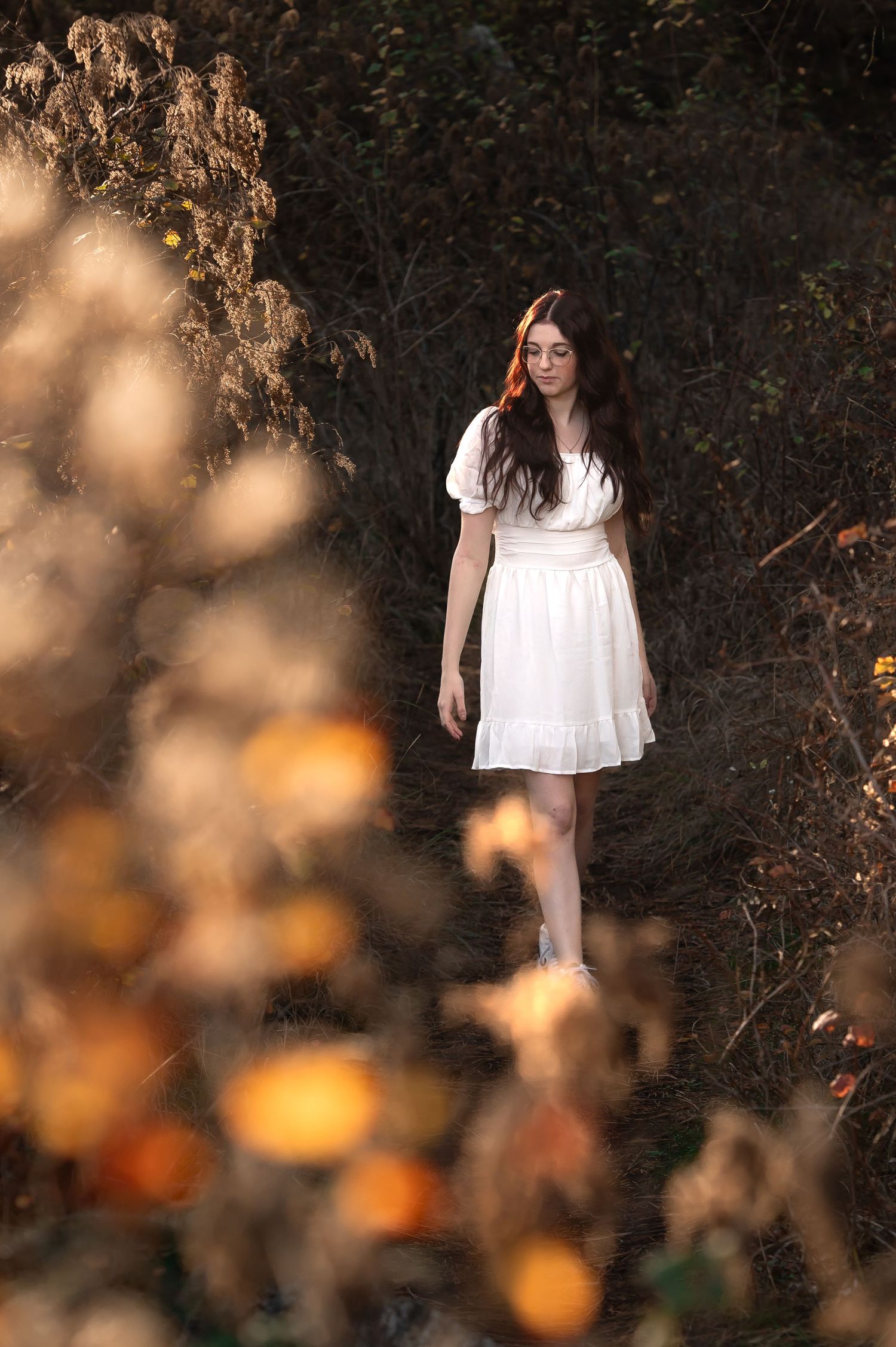 Young woman in white dress walking through autumn foliage in a dreamy outdoor nature setting.