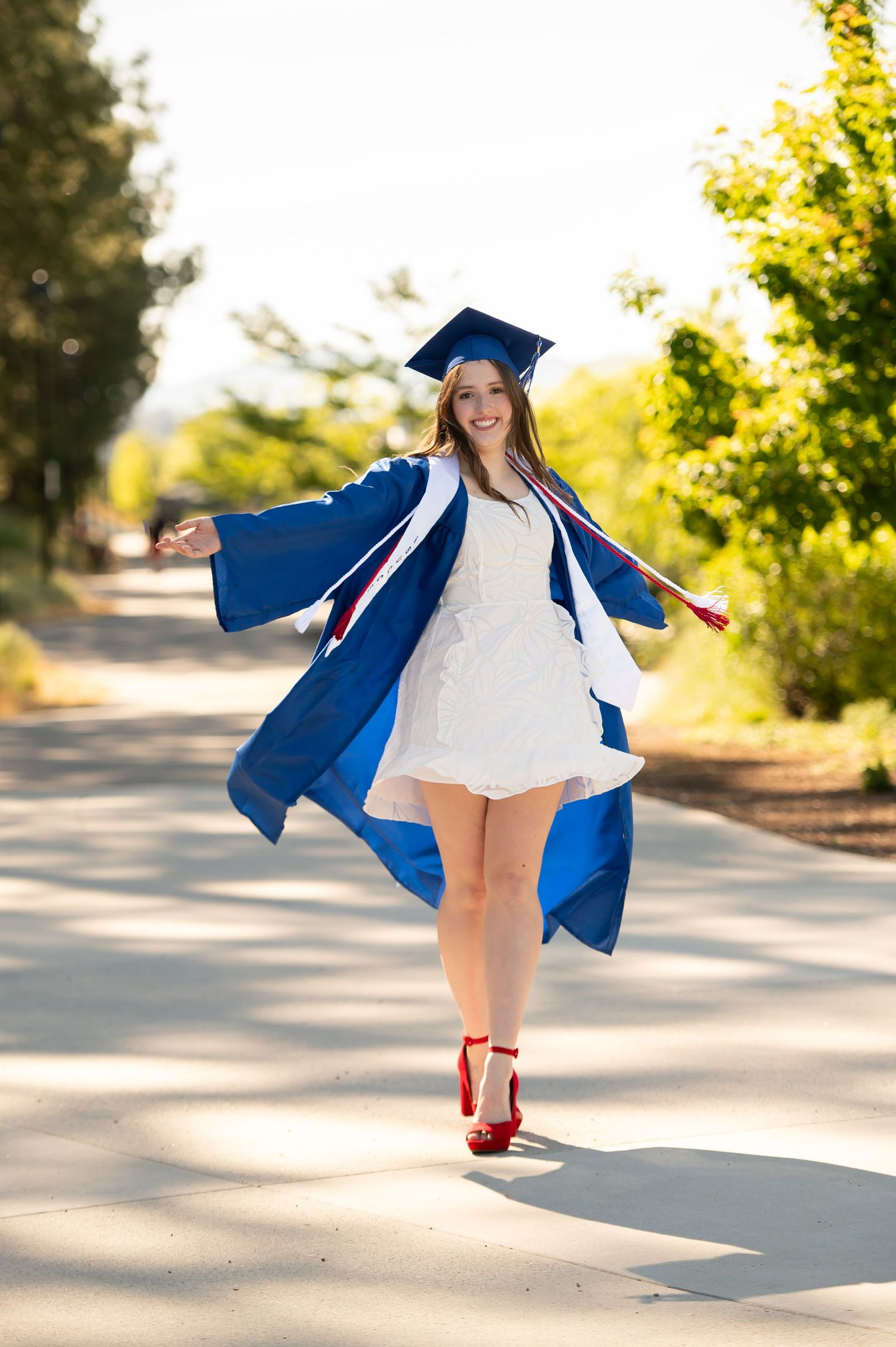 Happy graduate in blue cap and gown twirling on a sunny outdoor path.