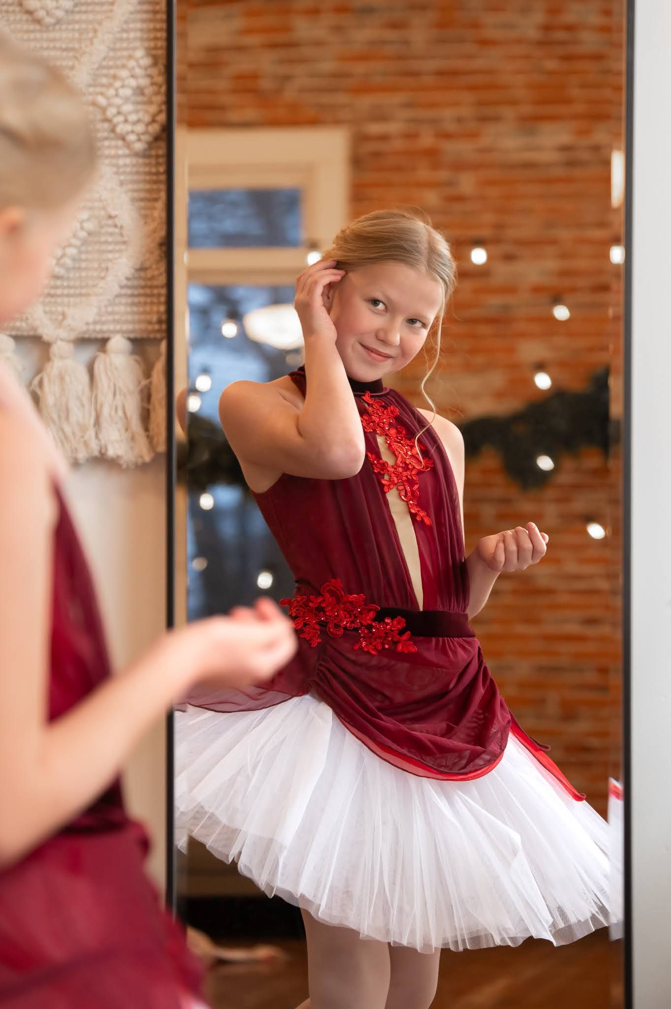 Young blonde girl in red white ballet tutu posing before mirror smiling.