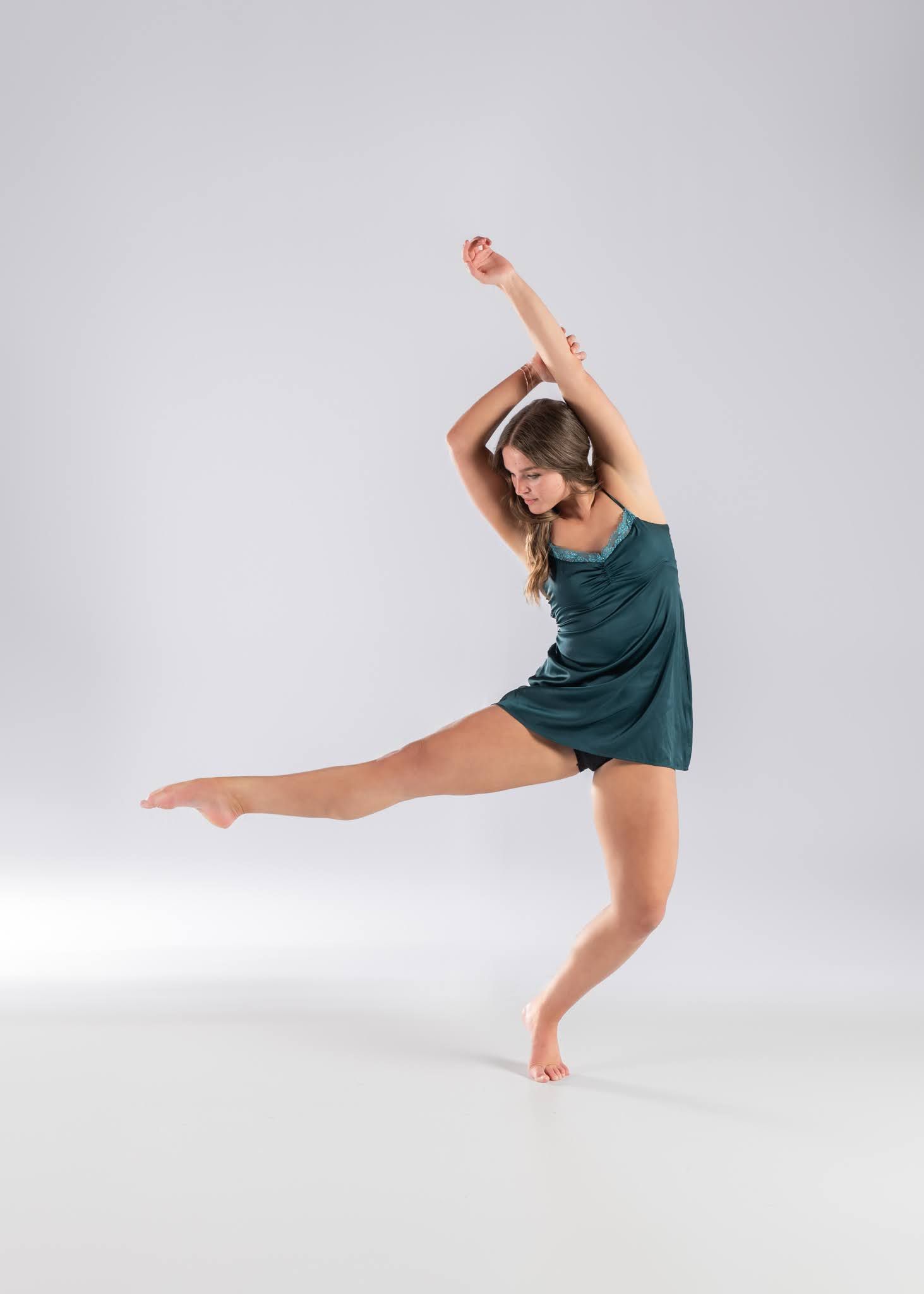 A dancer in a teal dress balances on one leg with arms raised in an elegant contemporary dance pose on a white background.