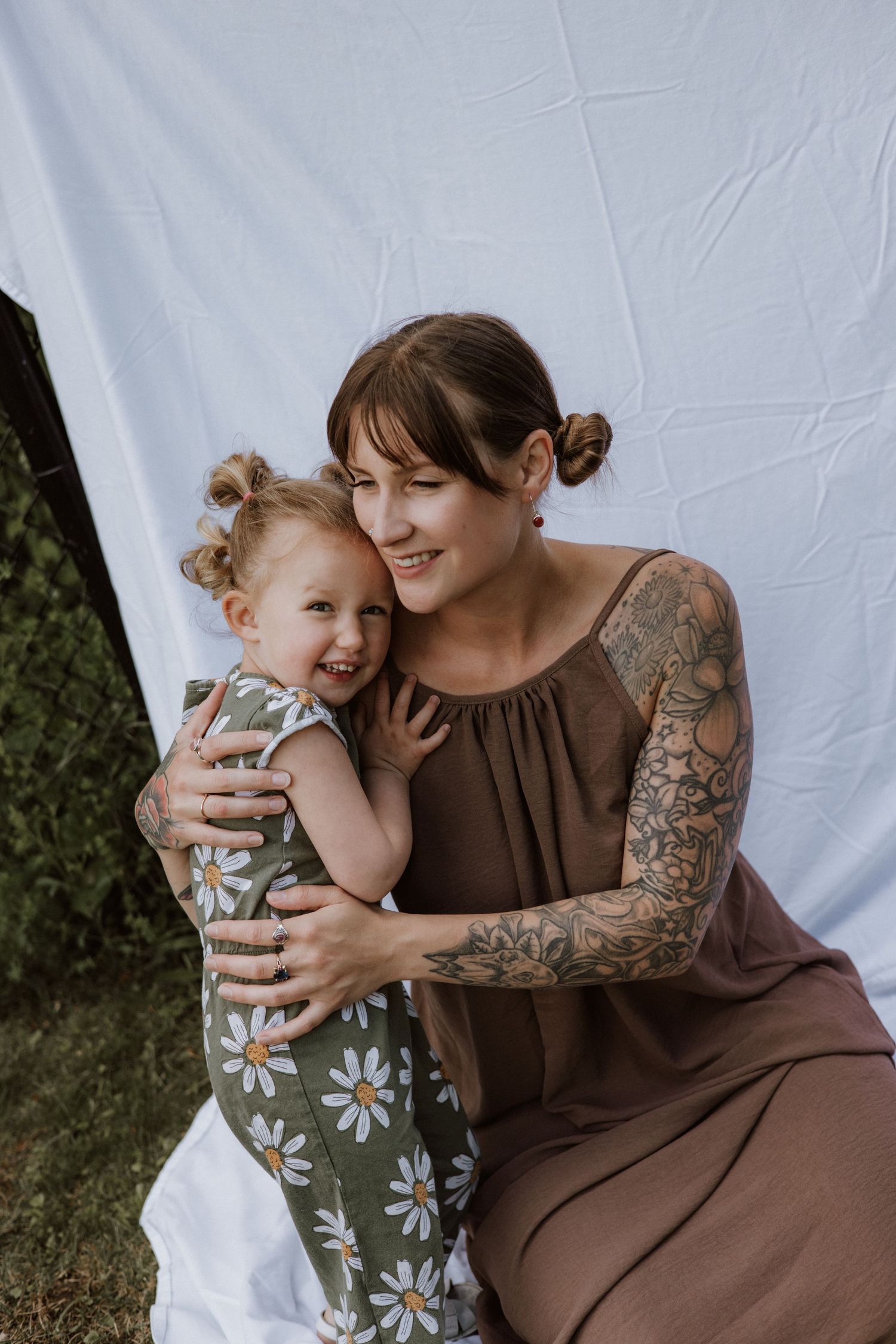 Tattooed mother in brown dress hugging laughing toddler girl in daisy outfit against white backdrop outdoors.