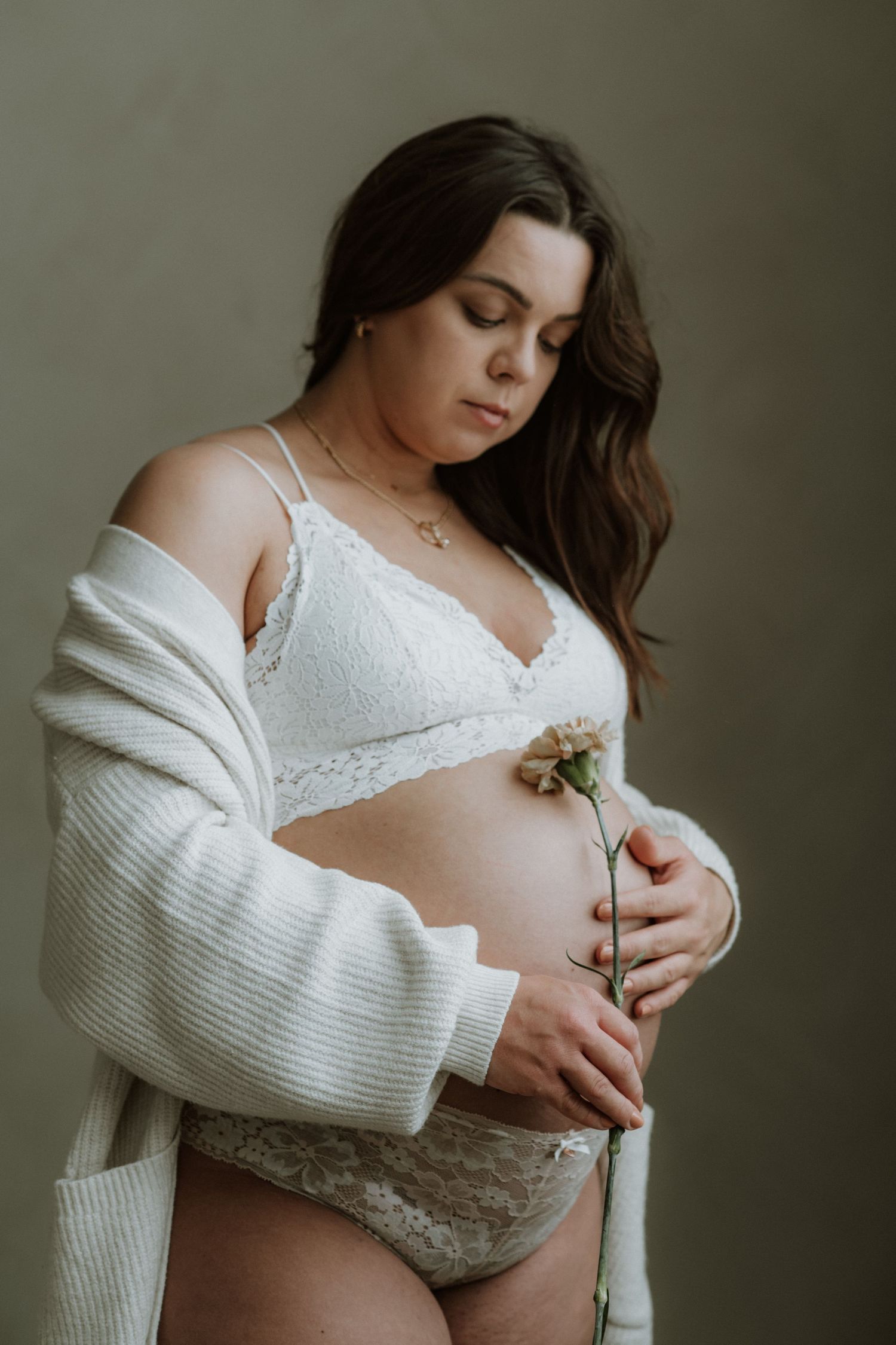 Pregnant woman in white lace lingerie and cardigan holds a flower against her baby bump in a studio.