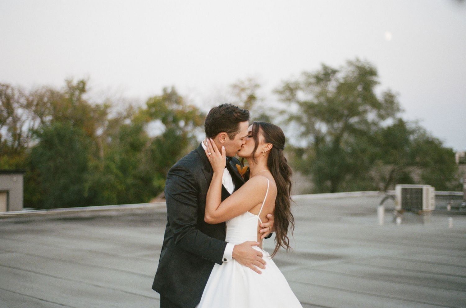 Bride and groom sharing a passionate kiss on a rooftop with trees in the background.