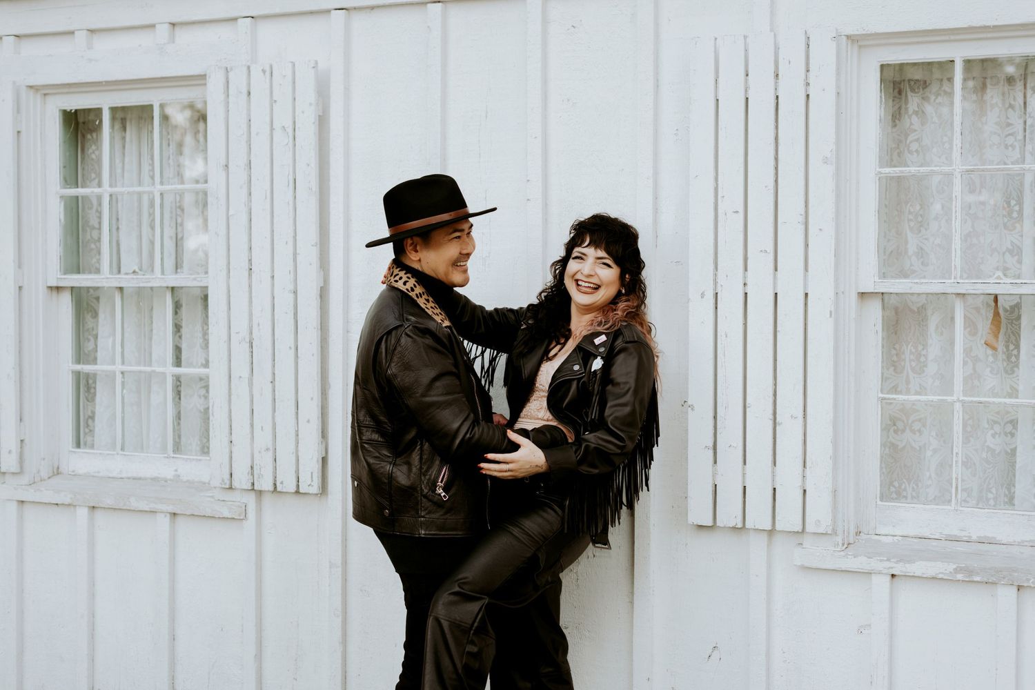 Joyful couple in leather jackets laughing together against a white wooden building exterior.