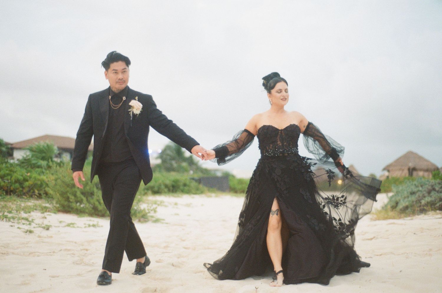 Bride and groom in black wedding outfits walk hand in hand on tropical beach.