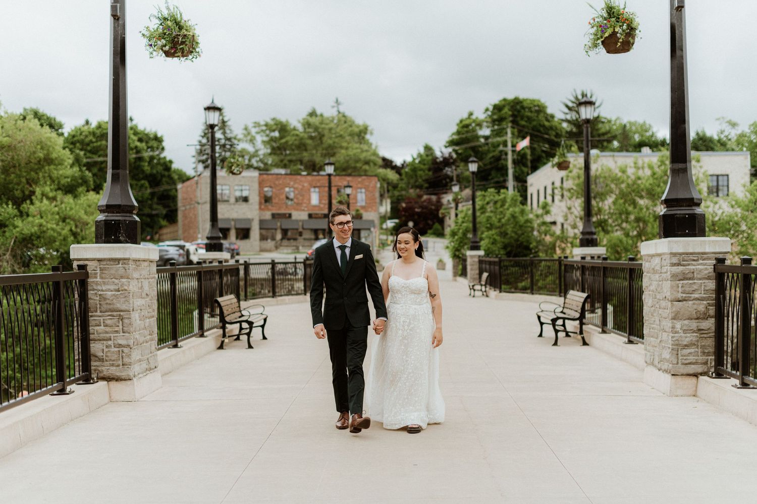 Bride and groom walking hand in hand across a stone bridge with hanging flower baskets.