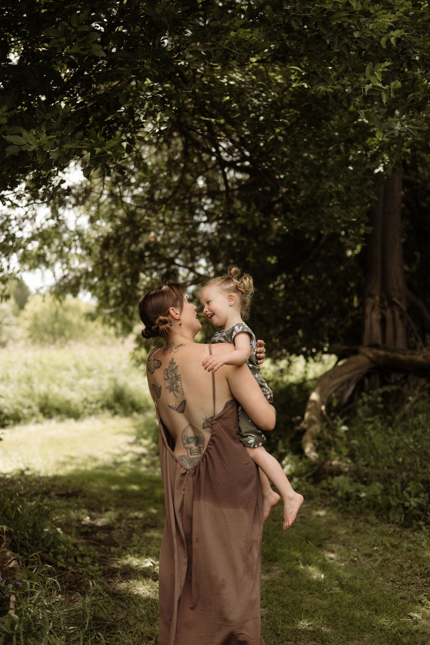 A tattooed mother in a brown dress holds her laughing toddler face-to-face under a leafy tree.