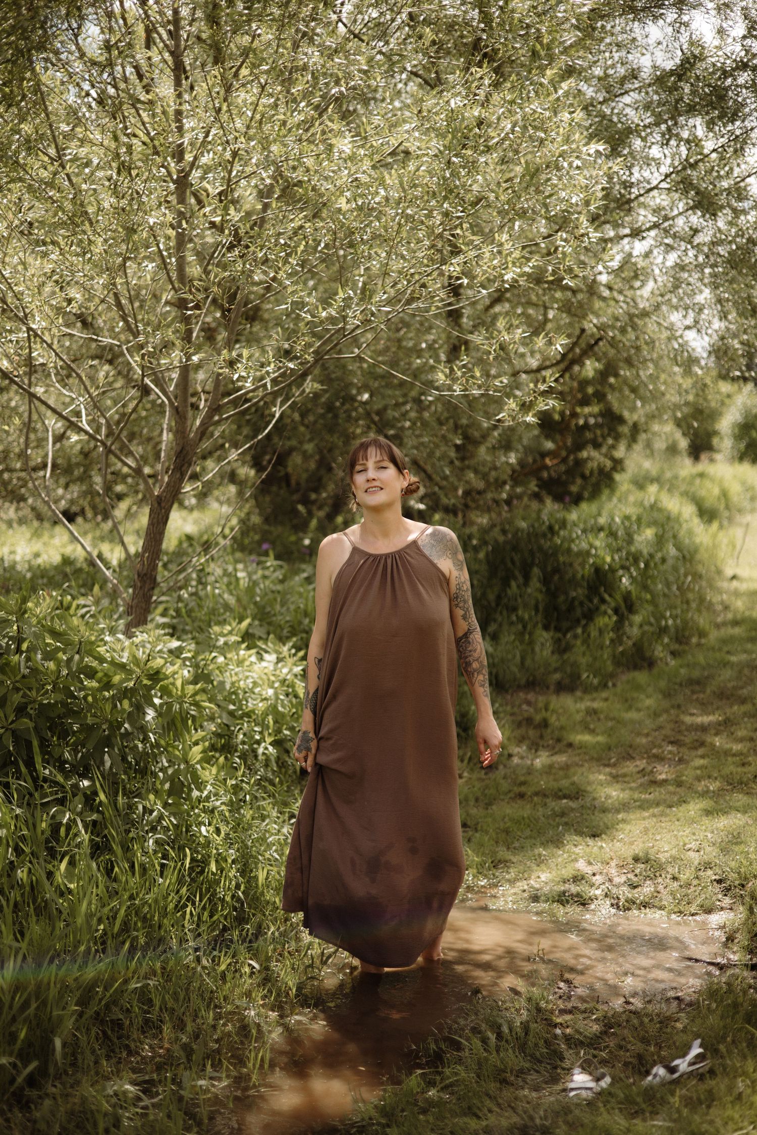Smiling tattooed woman in brown maxi dress stands in shallow stream surrounded by green trees.