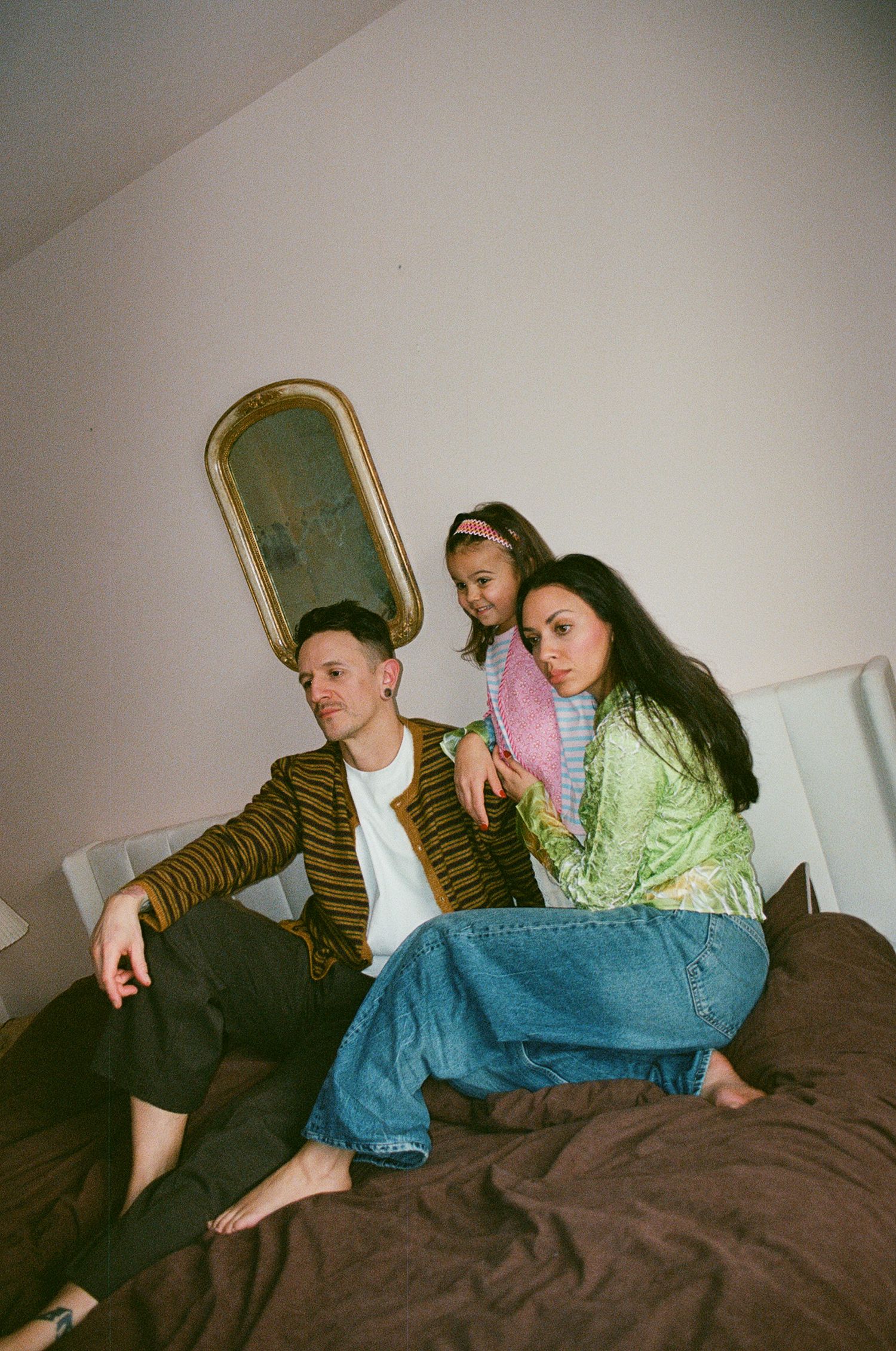 Family of three sits on bed, young girl in pink between parents, gold ornate mirror on wall above.