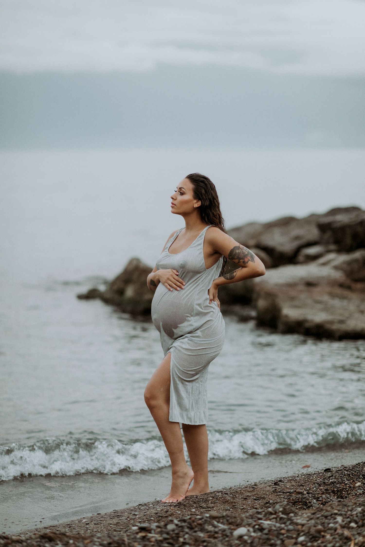 Pregnant tattooed woman in grey dress standing barefoot on rocky shoreline during moody maternity shoot.