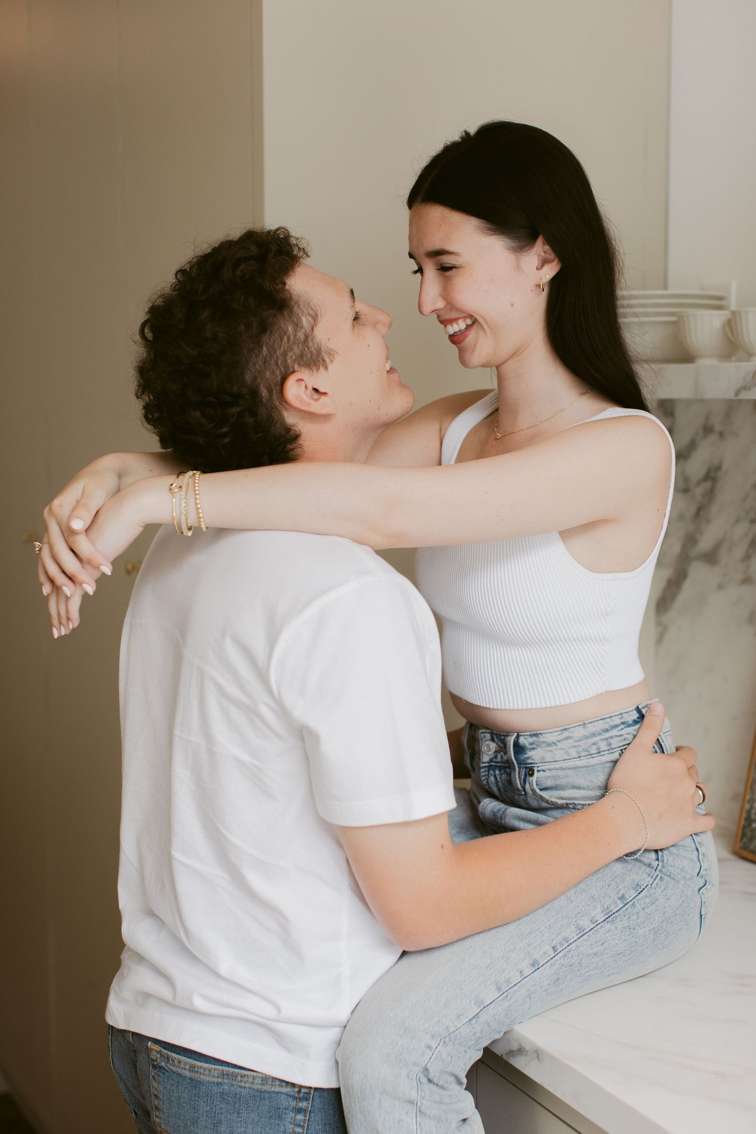 Smiling woman in white crop top sitting on counter embracing her partner in a bright kitchen.