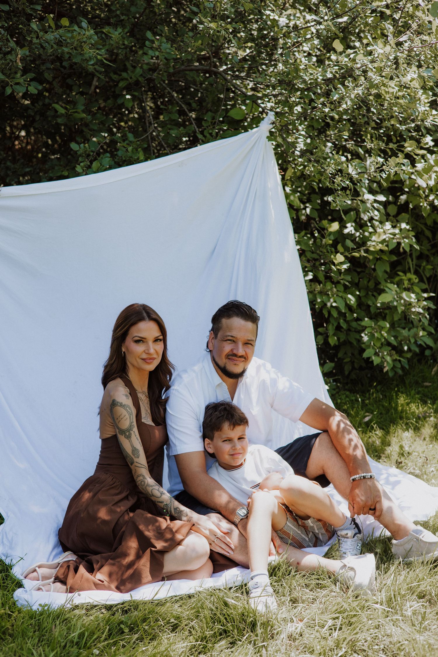 Family of three in white and brown outfits sit together on a picnic blanket in front of a white backdrop and green leafy bushes.