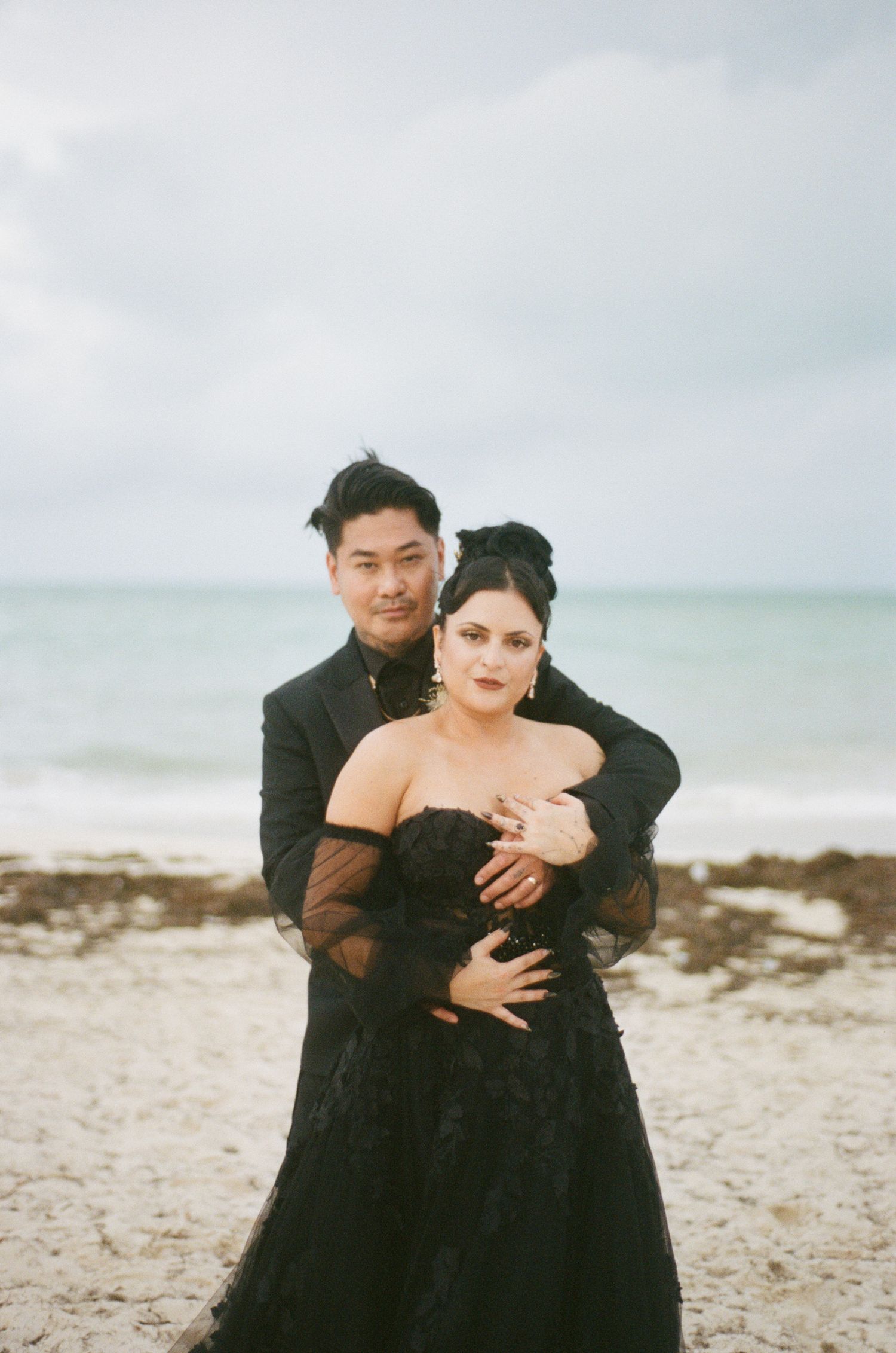 Elegant couple in black wedding outfits embracing on a sandy beach with ocean backdrop.