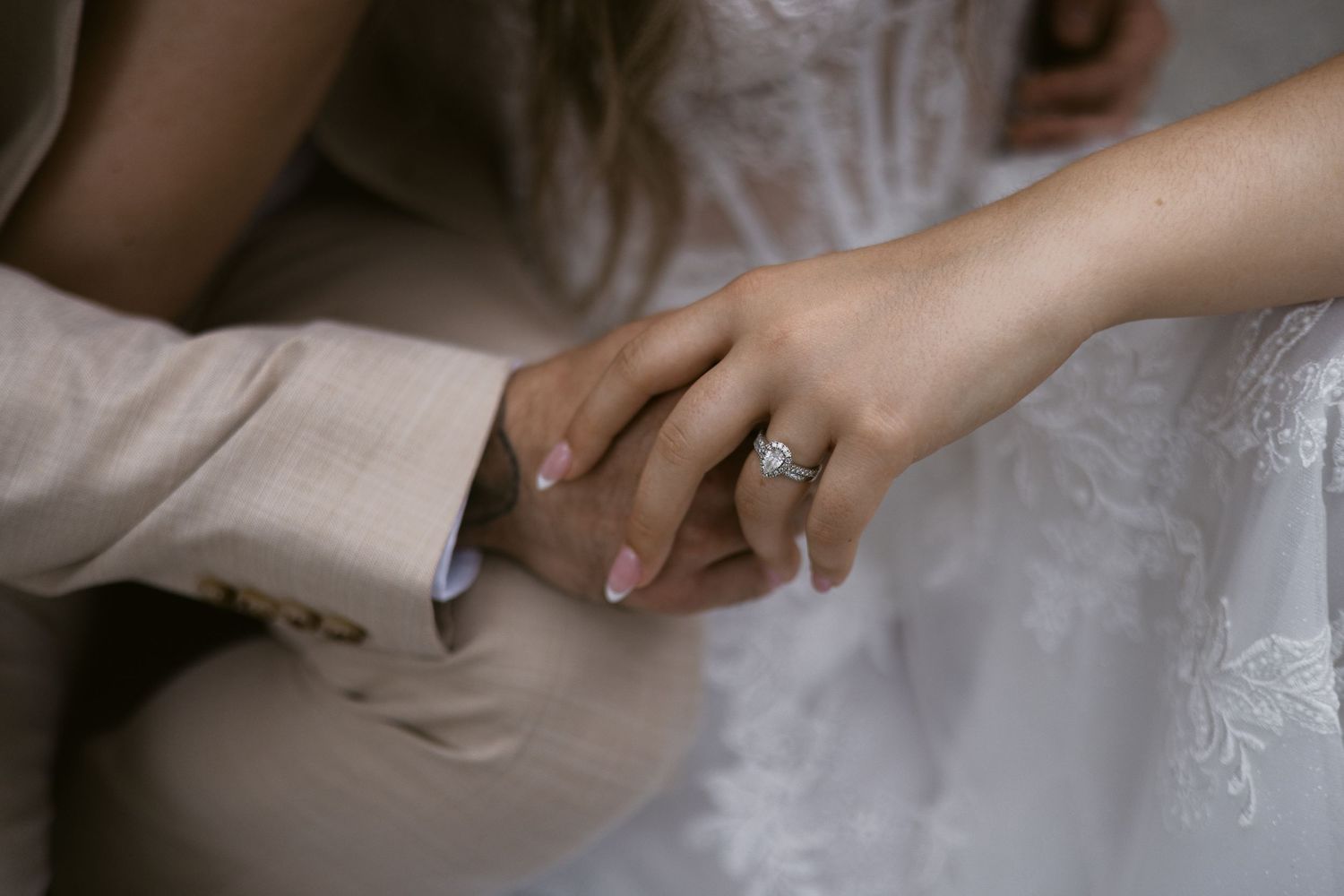Close-up of bride's hand with diamond engagement ring resting on groom's hand in beige suit.