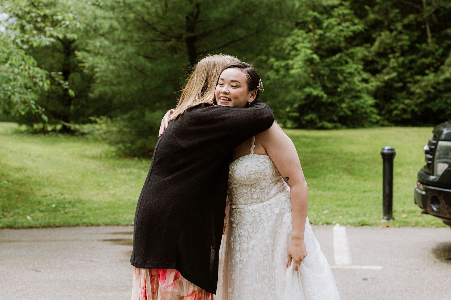 Bride in lace wedding gown shares a warm hug with a female guest outdoors at wedding venue.