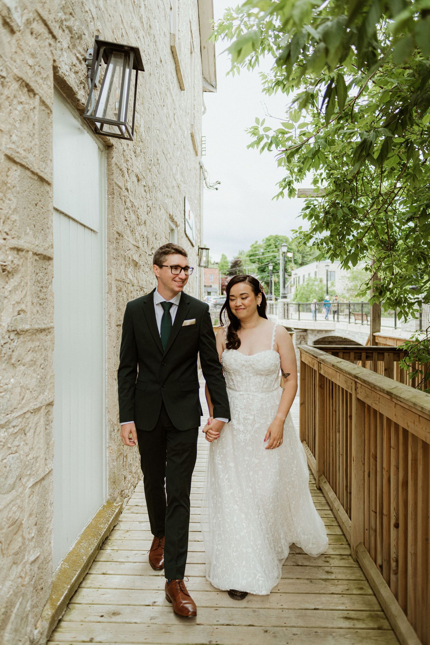 Smiling groom in black suit and bride in lace wedding gown walk hand in hand along outdoor walkway.