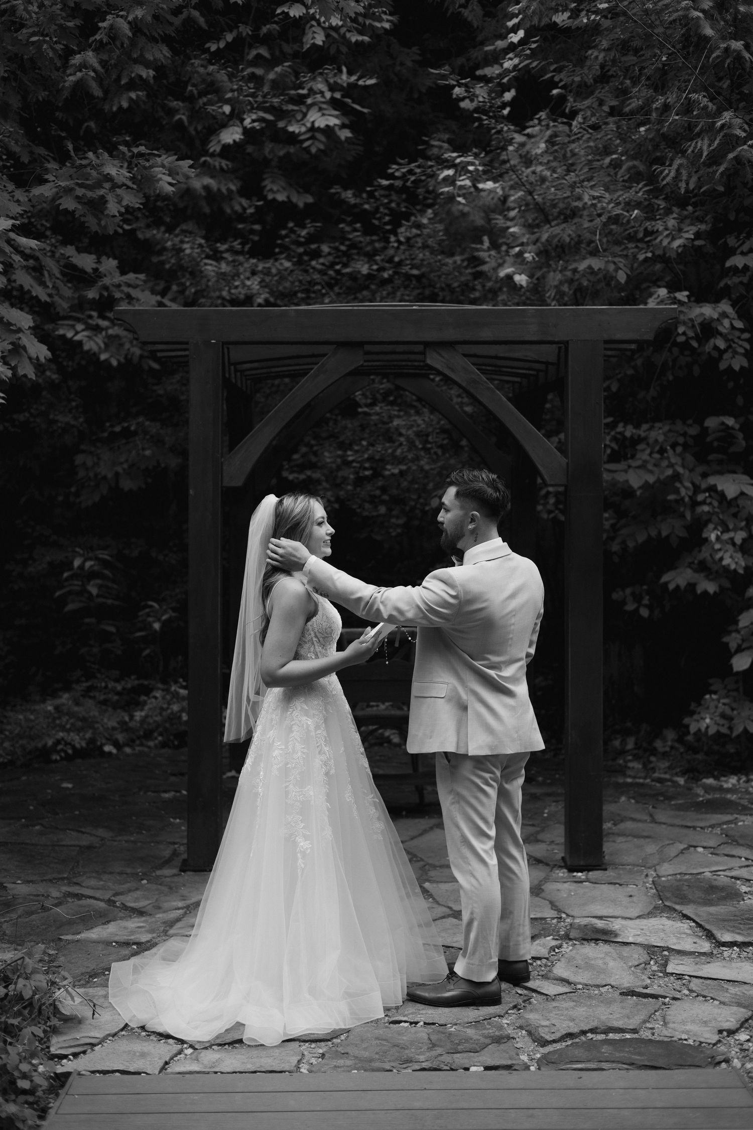 Black and white photo of groom lifting brides veil under wooden arch after outdoor ceremony.