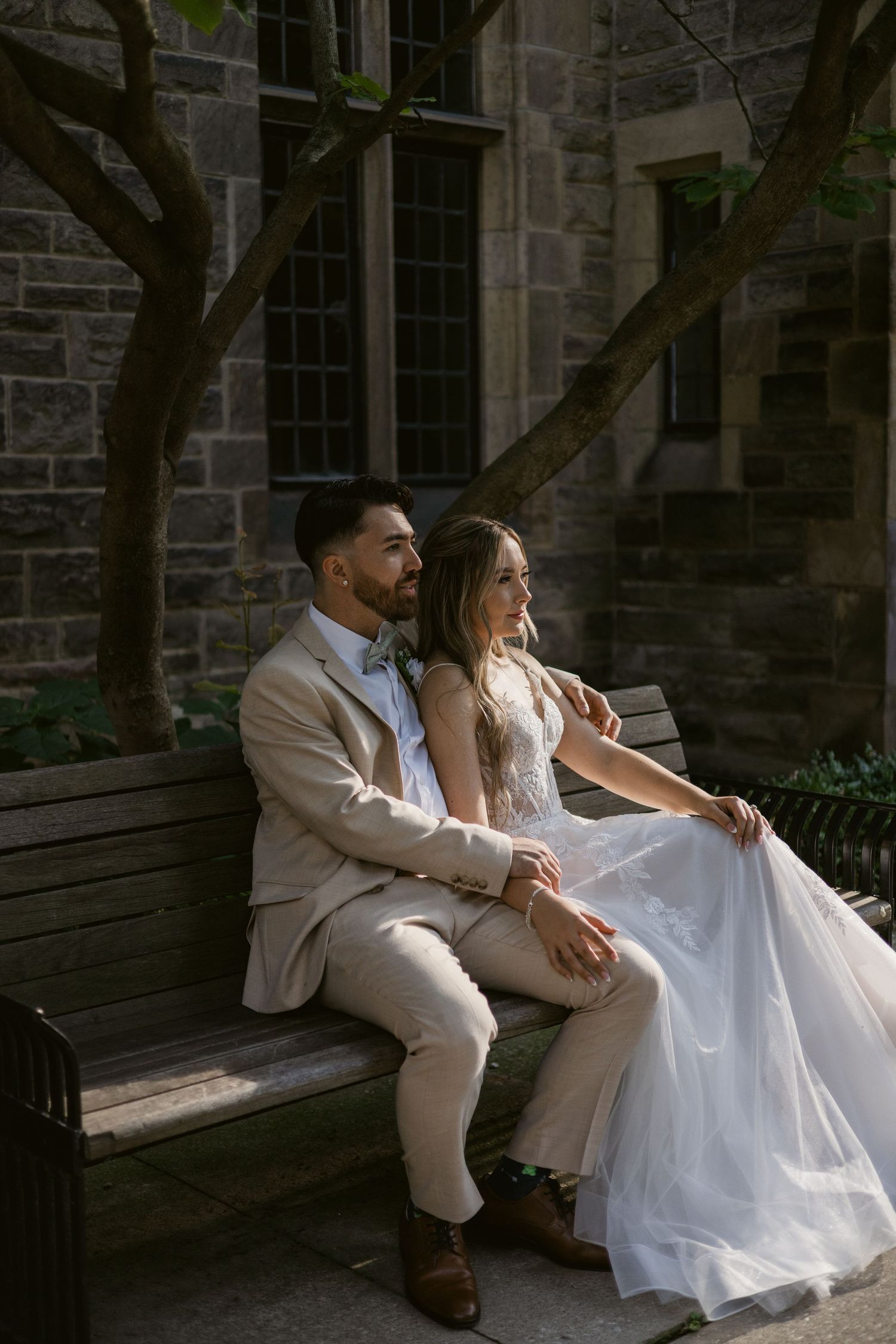 Bride and groom sit closely together on a wooden bench in dappled sunlight beside a historic stone building.