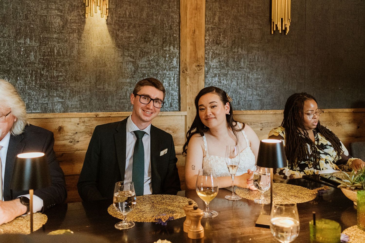 Bride and groom smiling together at wedding reception dinner table with wine glasses.