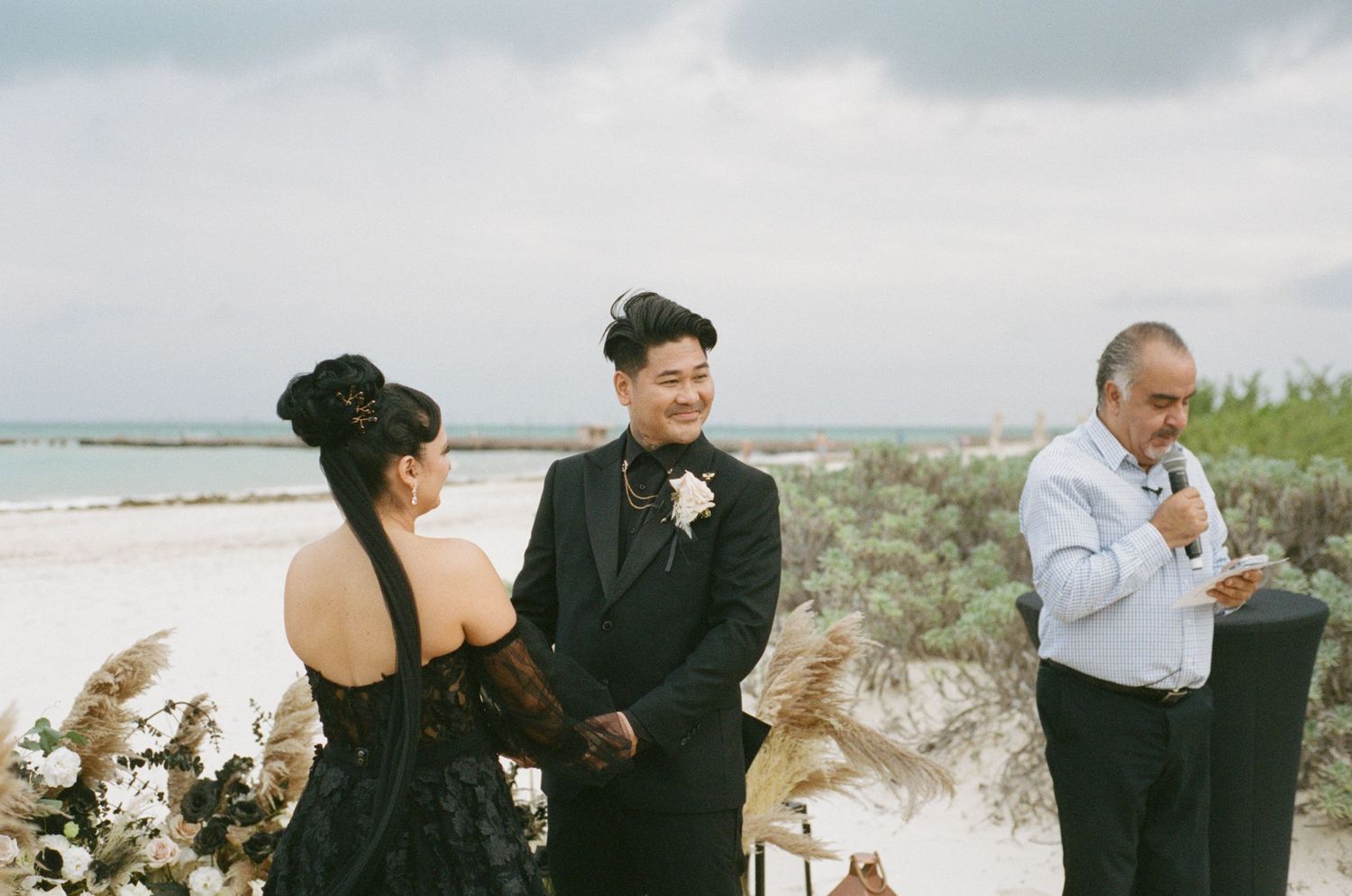 Groom smiling during beach wedding ceremony officiated by man in white shirt, with pampas grass floral arrangements.
