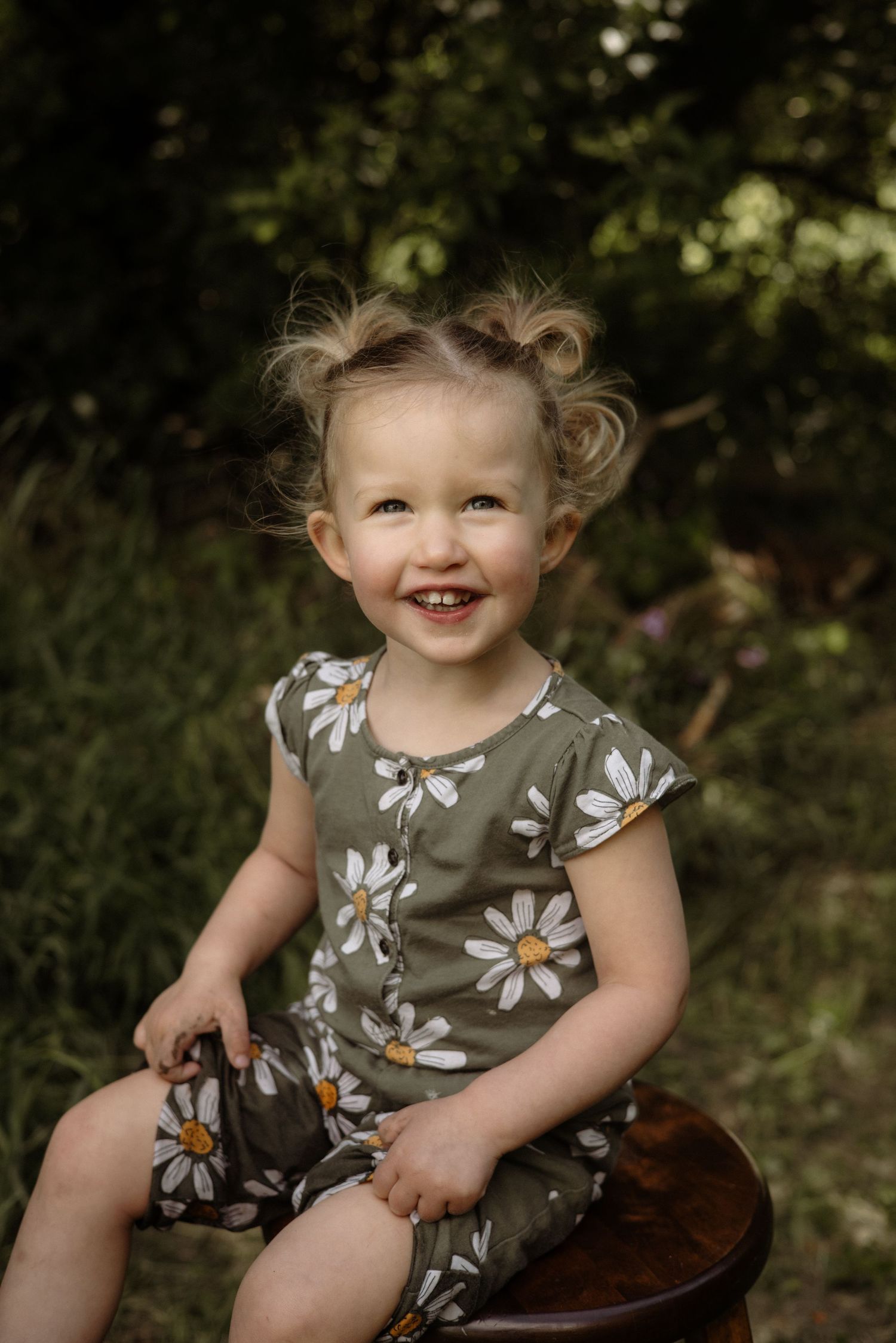 Cheerful toddler girl with pigtail buns smiling broadly while seated on wooden stool in daisy print romper outdoors.