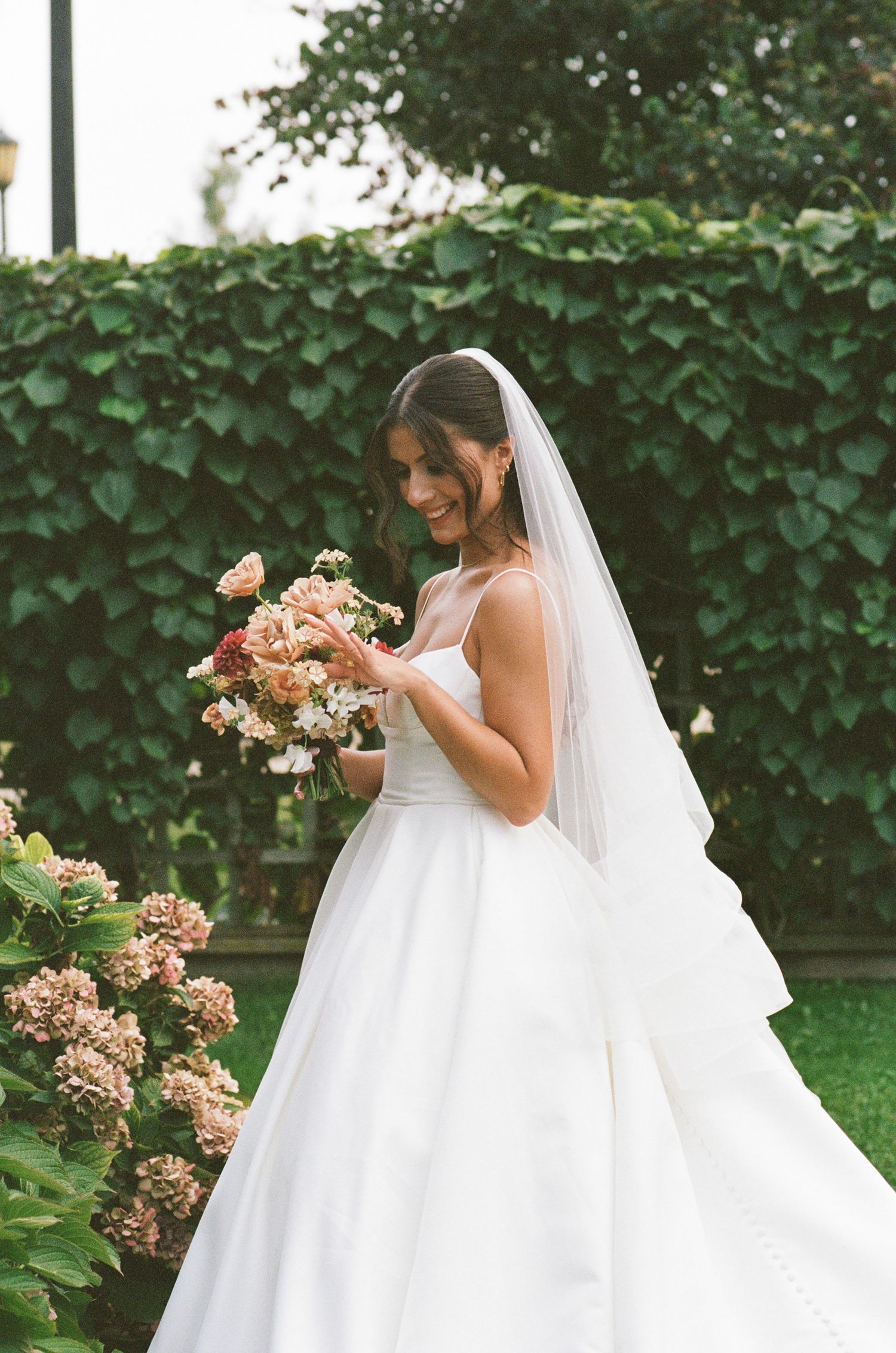 Smiling bride in a white ballgown and veil admiring her bridal bouquet in a lush garden setting.