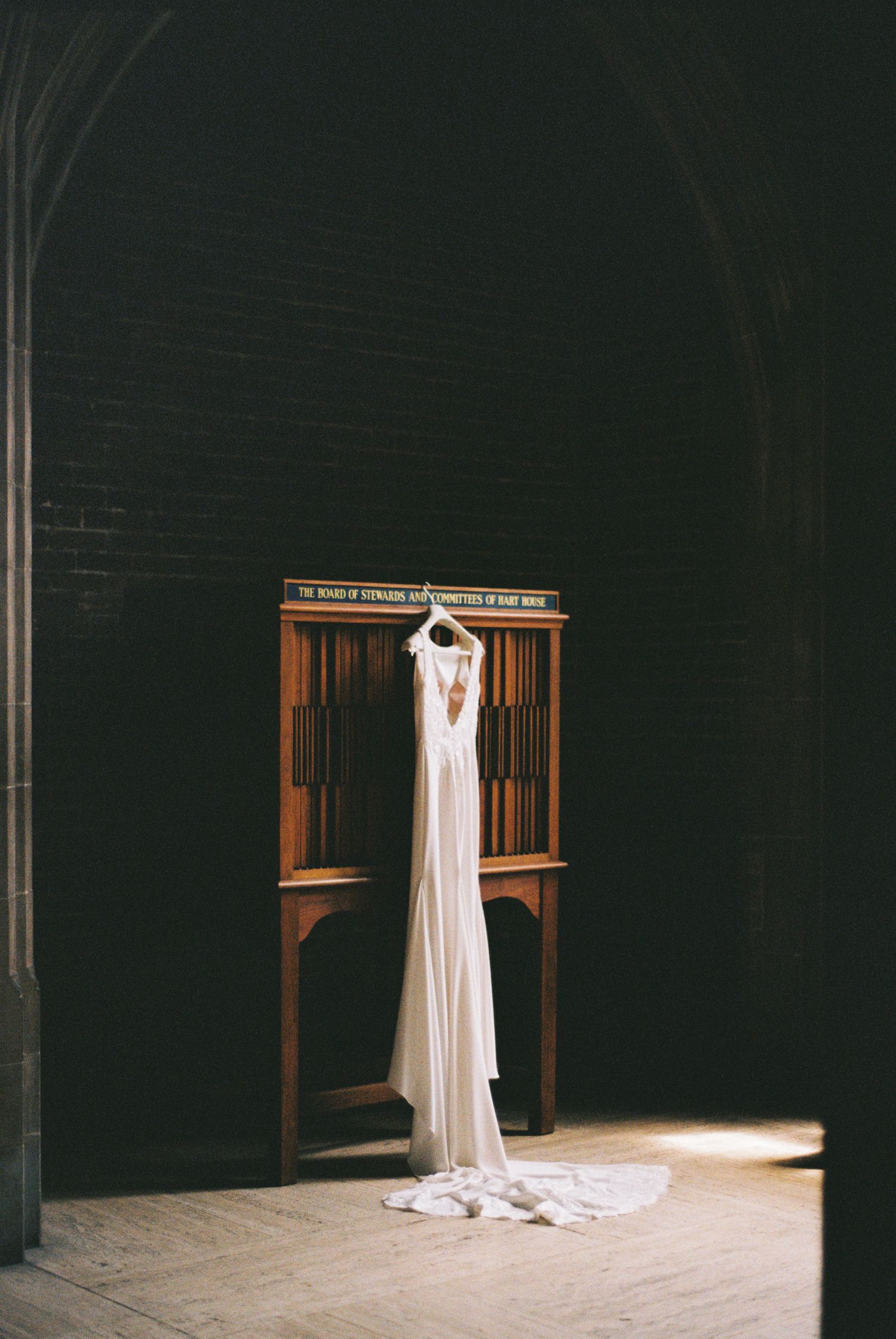 Elegant white wedding dress hanging on a wooden cabinet in a dimly lit stone church with arched ceilings.