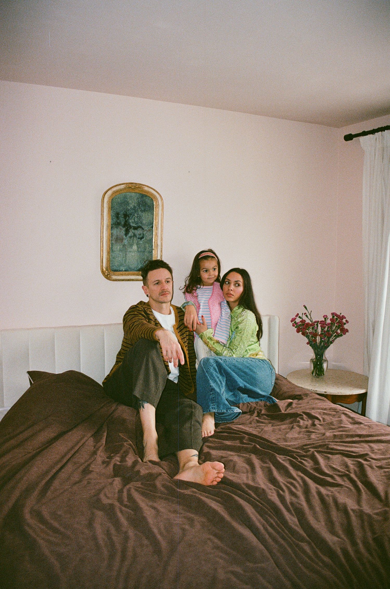 Family portrait on brown bed with gold mirror, man in cardigan, woman in green top holding young daughter.