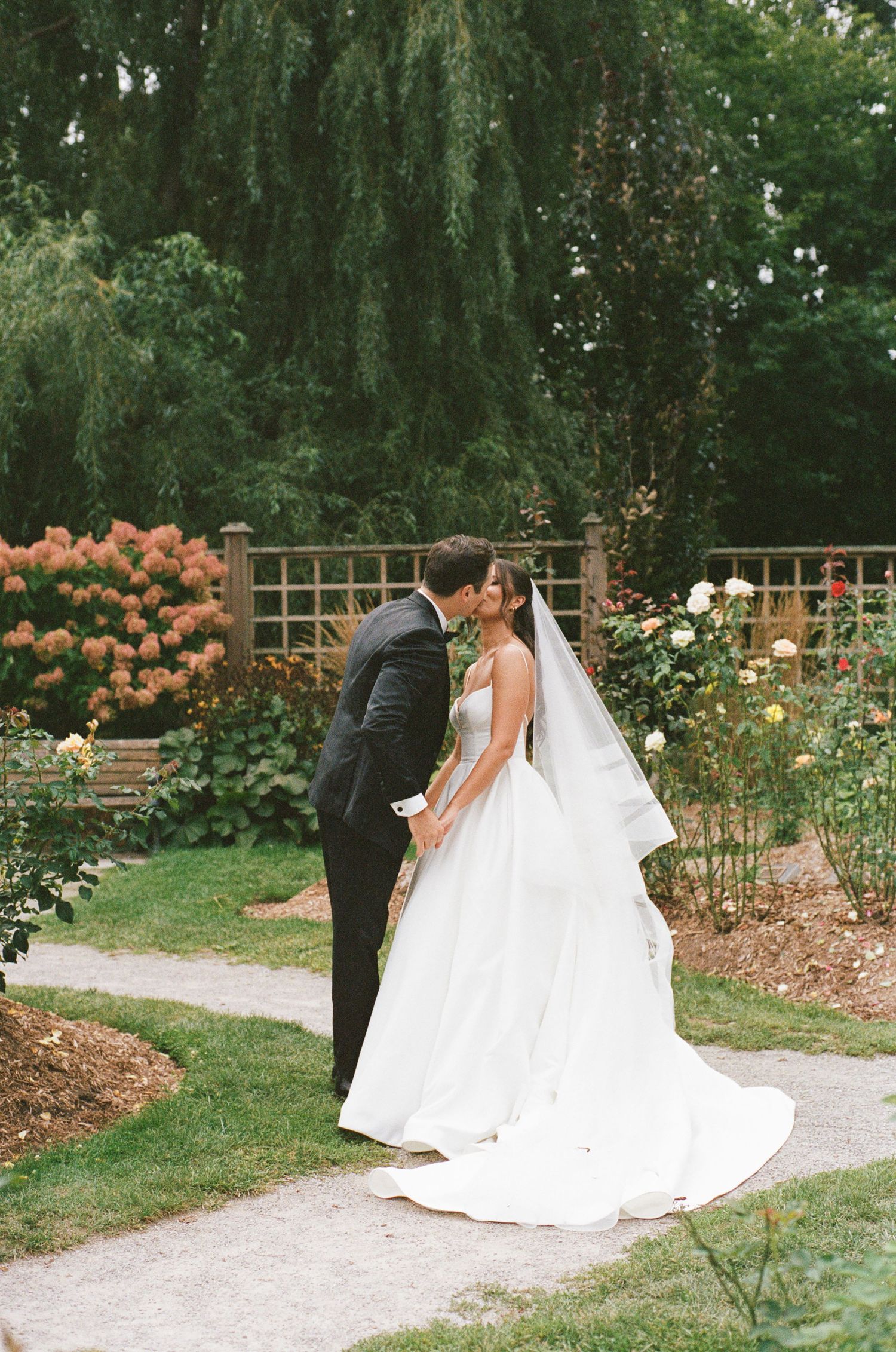 Bride and groom kissing in a lush garden surrounded by colorful roses and greenery.