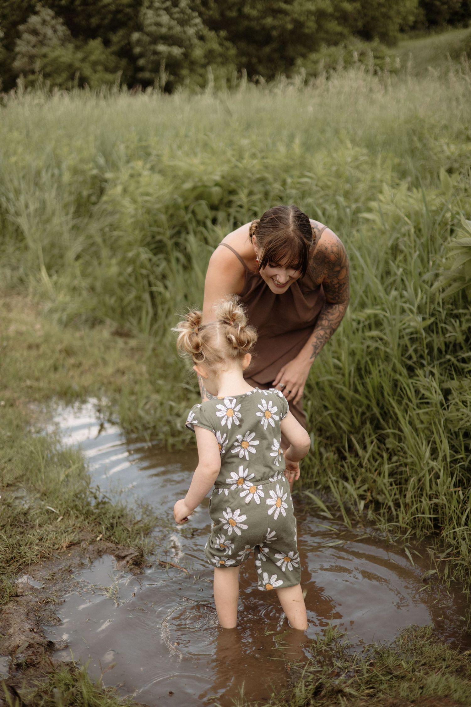 Mother with tattoos helps toddler girl in daisy romper wade through muddy puddle in tall grass.