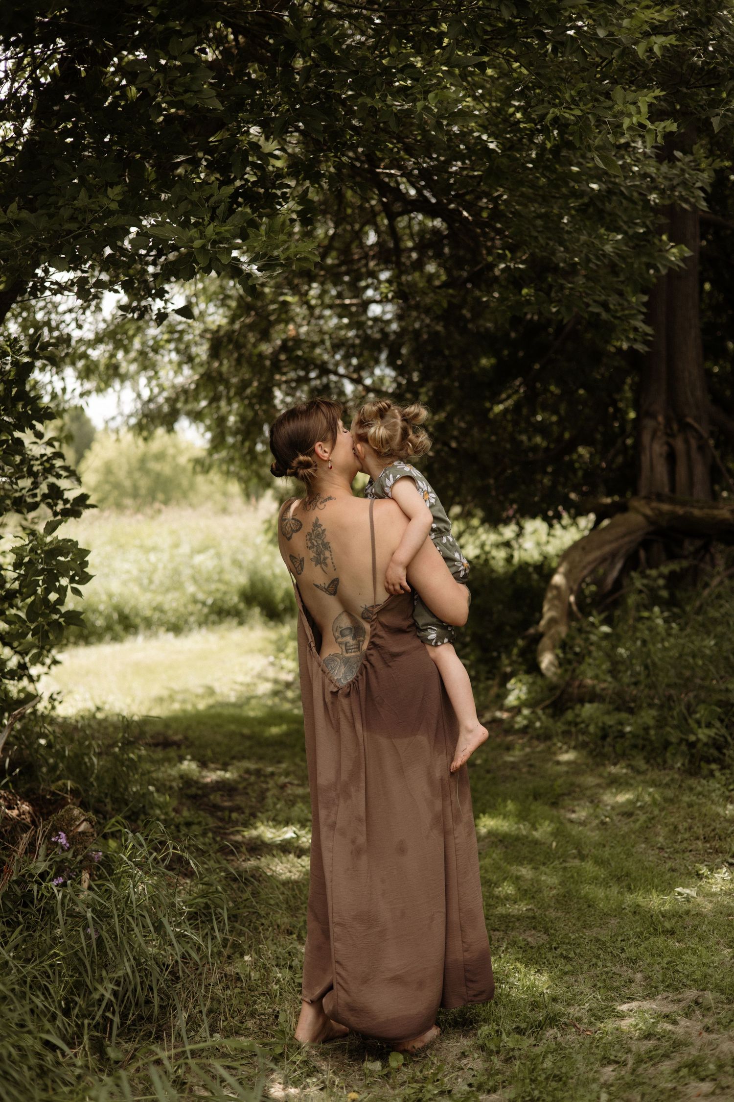 A tattooed mother in a brown backless dress holds and kisses her toddler child under leafy trees.