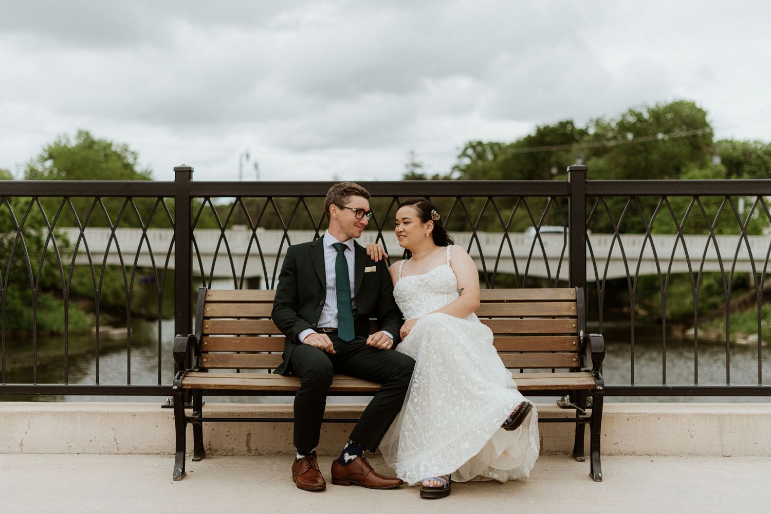 Bride and groom sit on a bench by a river, smiling at each other in wedding attire.