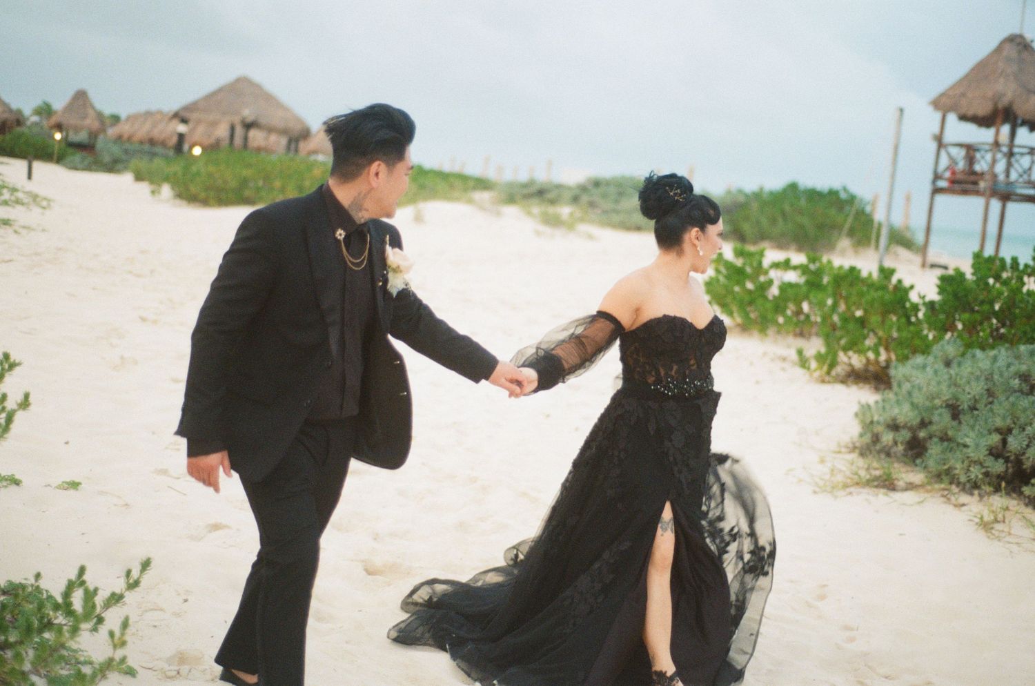 Groom in black suit holds brides hand in flowing black gown on sandy beach.