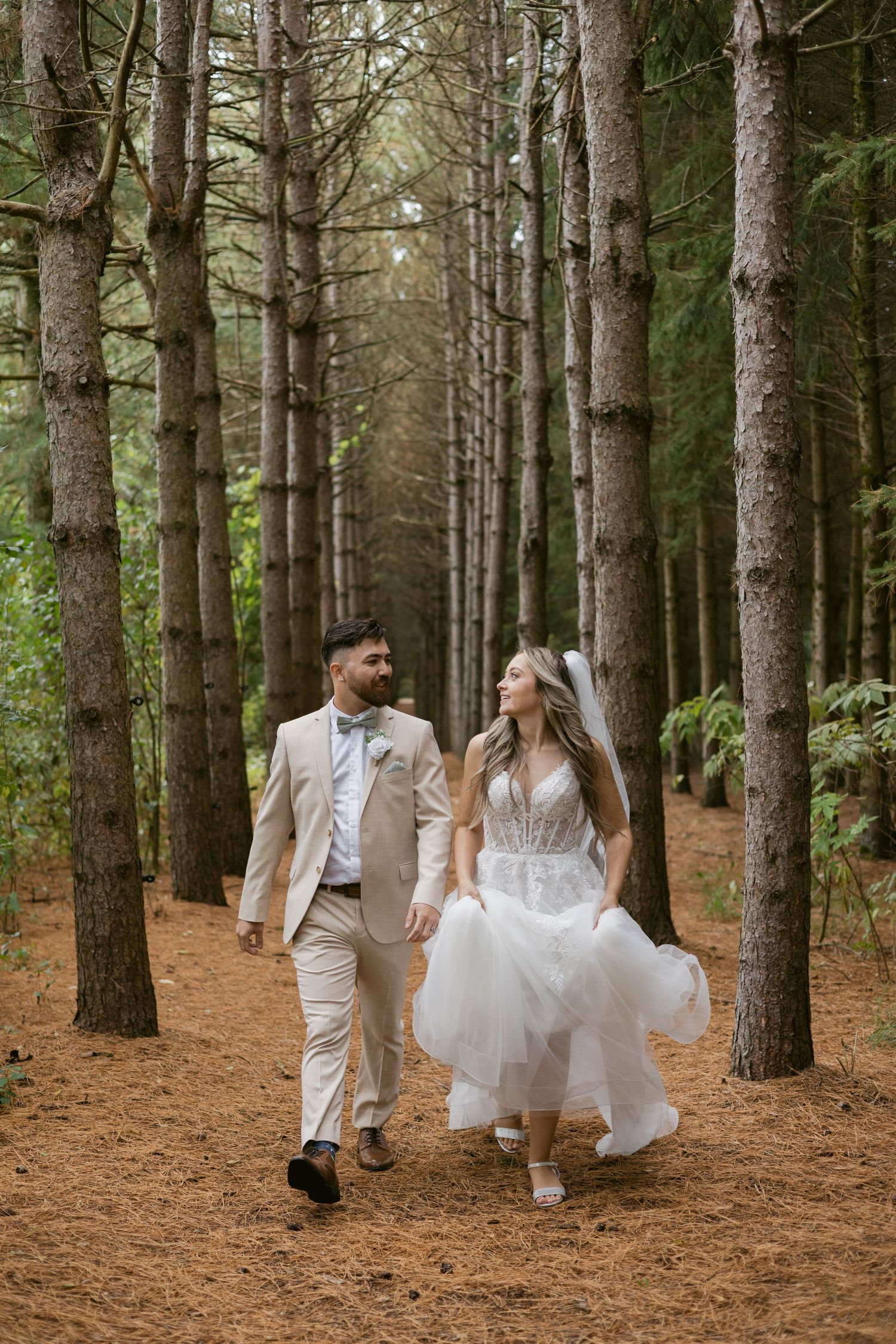 Smiling bride and groom walk through a pine forest, bride in lace gown holding her skirt.
