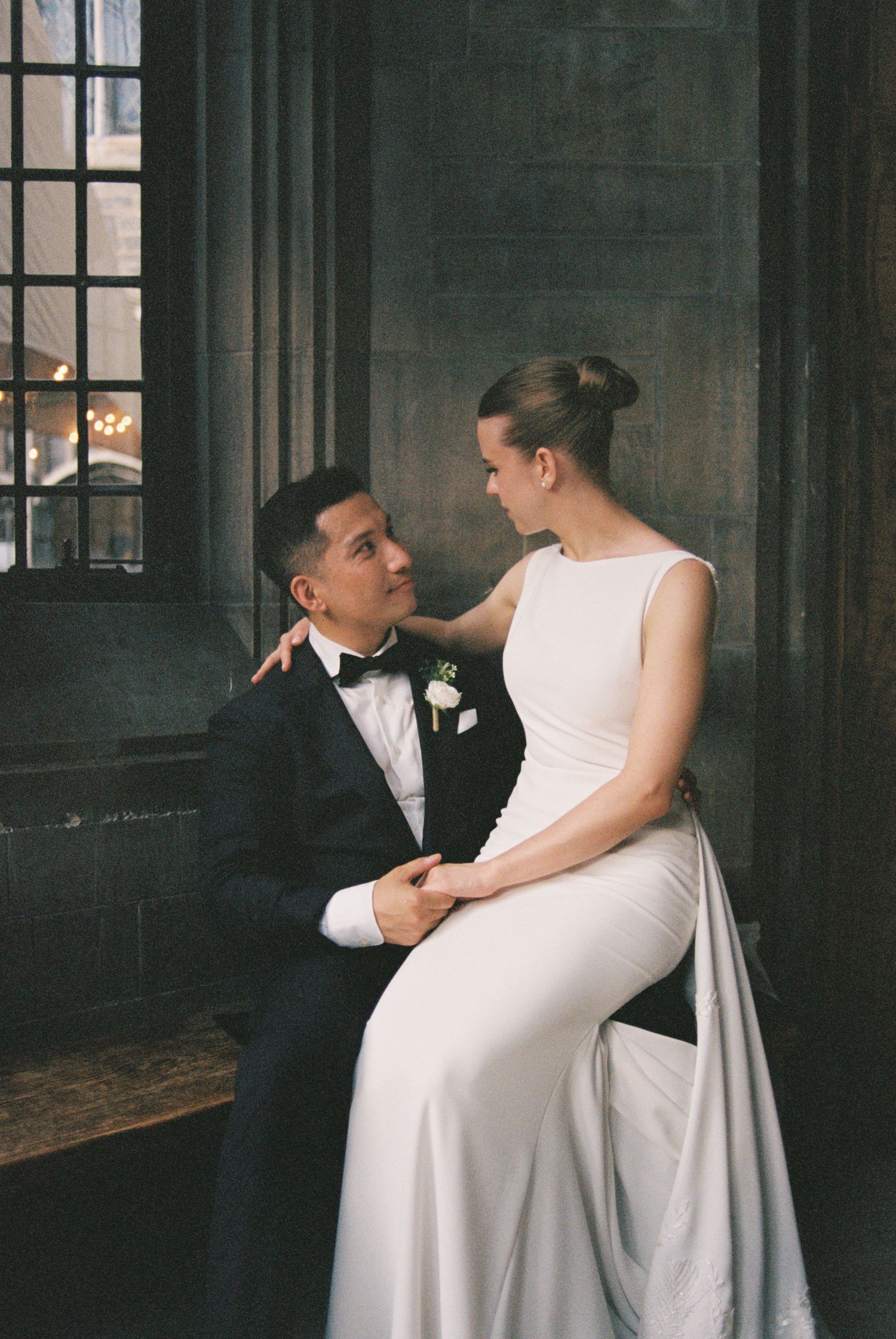 Elegant bride in white gown sits with groom in black tuxedo by gothic window in romantic wedding portrait.