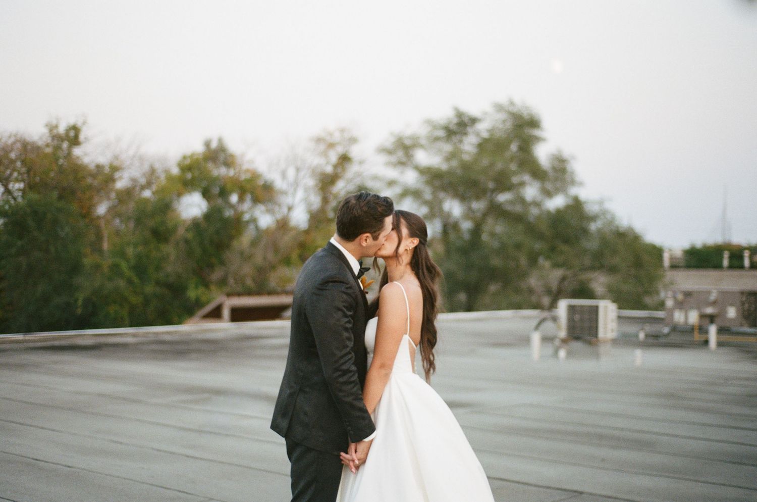 Bride and groom share a romantic rooftop kiss in elegant wedding attire against a soft sky backdrop.