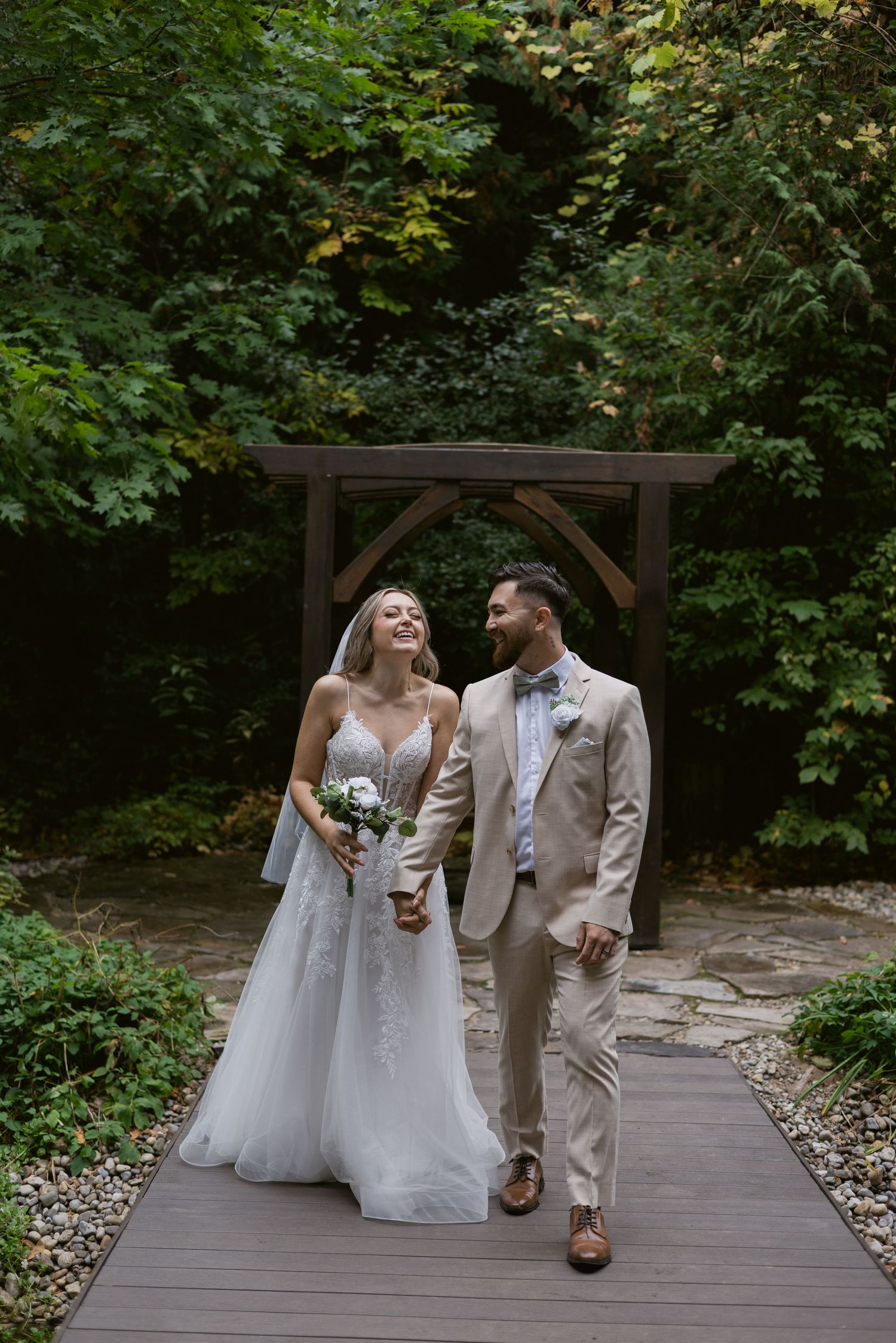 Bride and groom walking hand in hand on wooden bridge path with lush green trees behind.