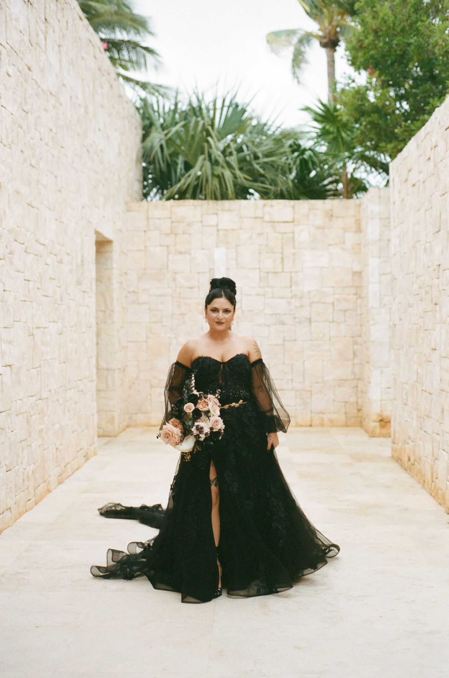 Bride in dramatic black lace wedding dress with sheer sleeves posing in stone courtyard.