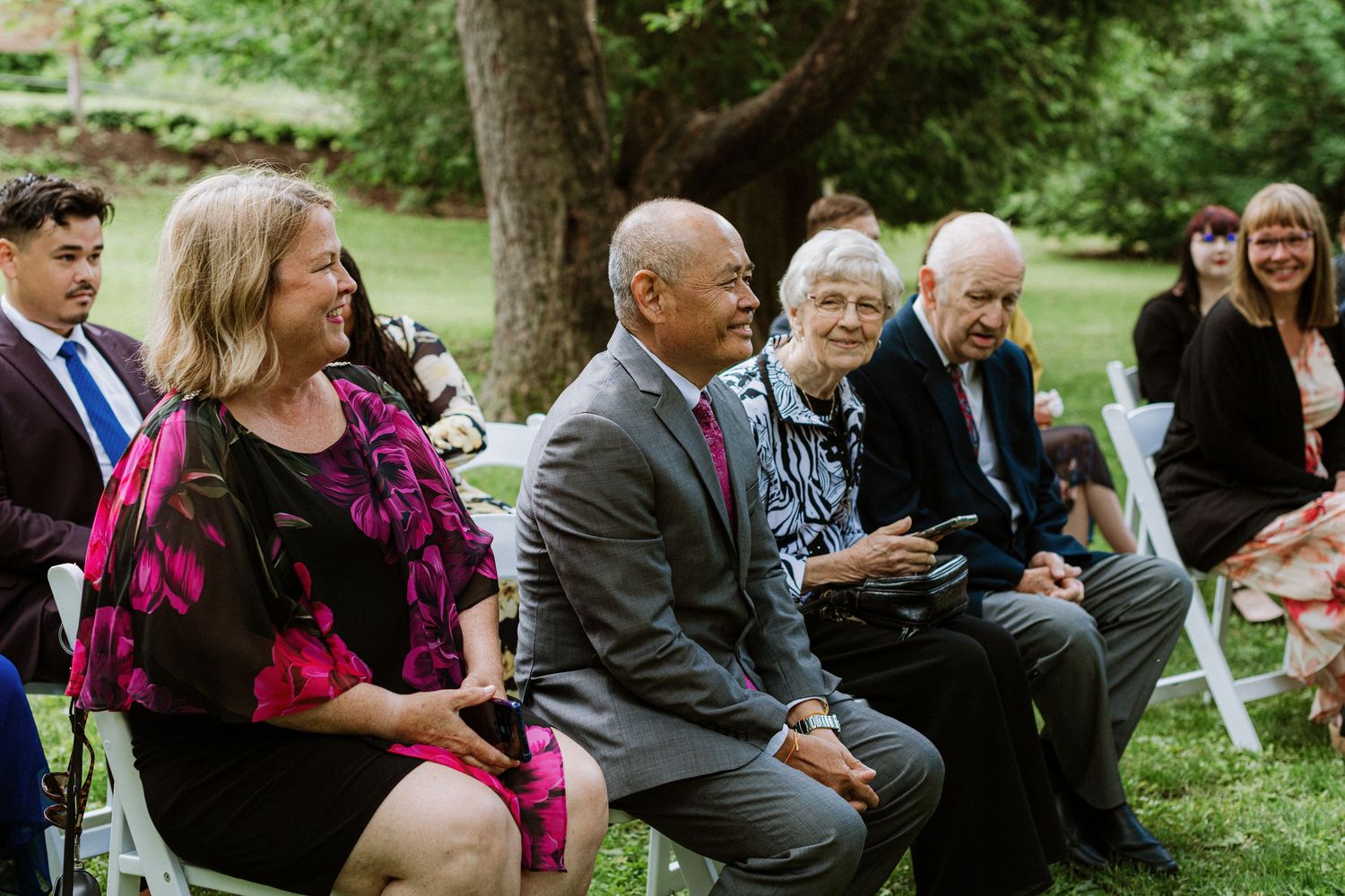 Wedding guests smile and watch outdoor ceremony seated on white chairs in lush green garden setting.