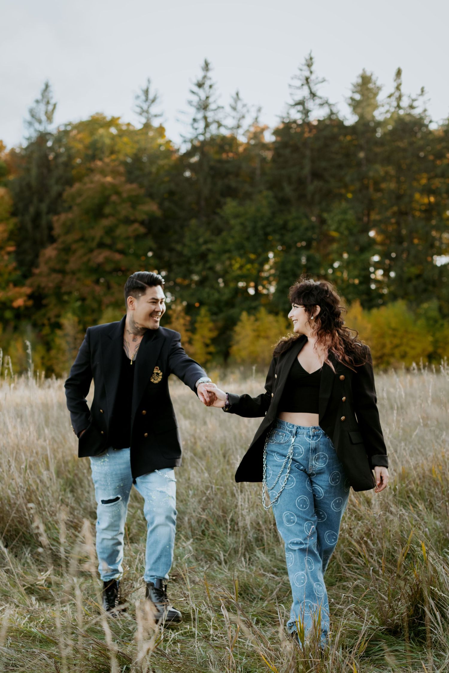 Smiling couple in matching black blazers and jeans holding hands while walking through golden autumn grass field.
