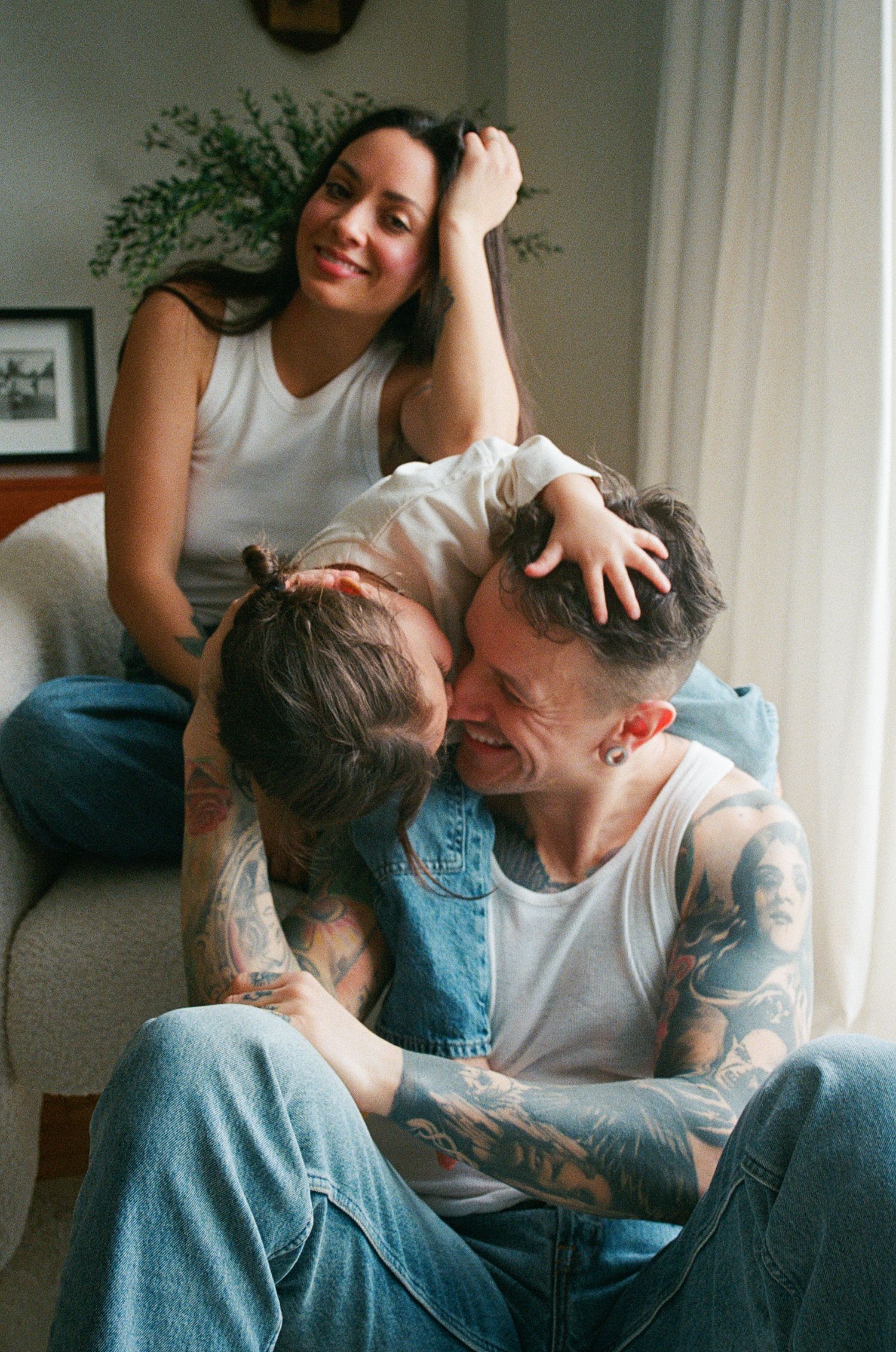 Smiling tattooed couple with child sharing a joyful embrace near a bright window.