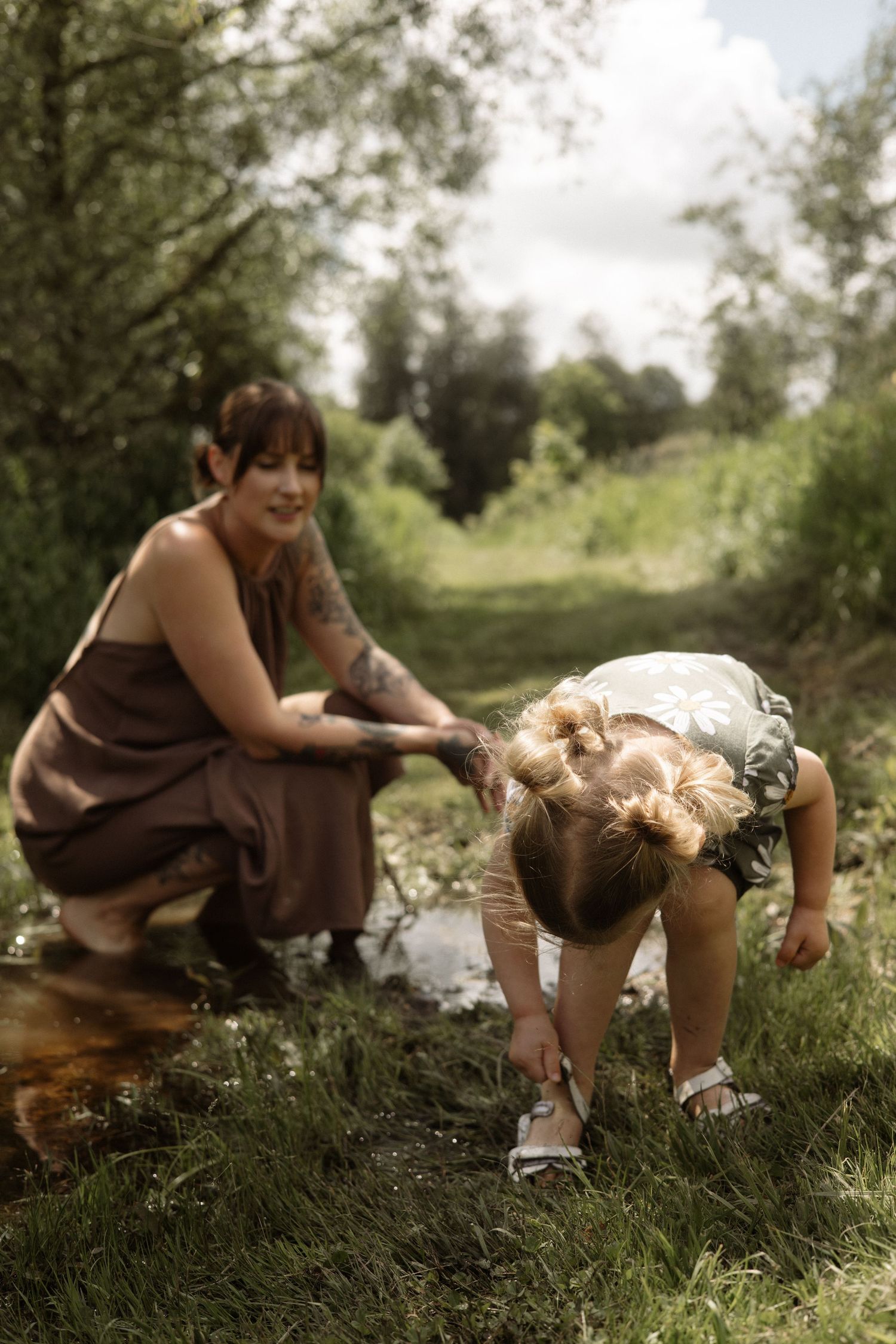 Tattooed mother watches smiling as toddler daughter bends down to remove sandals beside a small stream in green nature.
