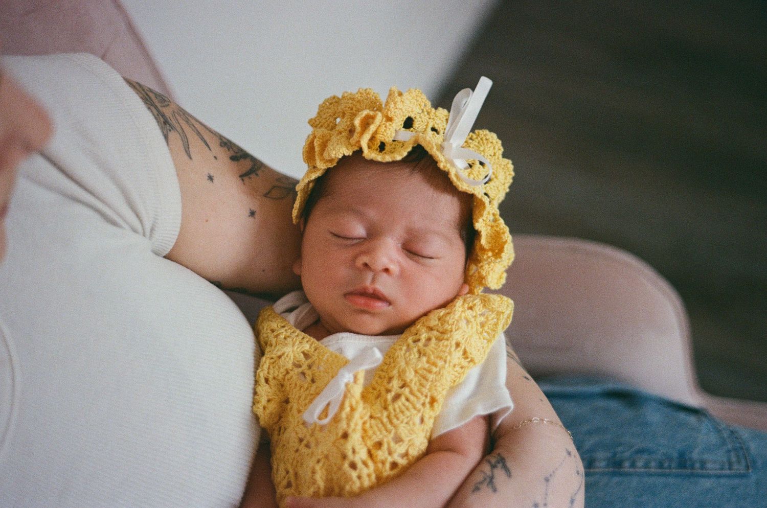 Newborn baby in yellow crocheted outfit sleeping peacefully in a tattooed mothers arms.