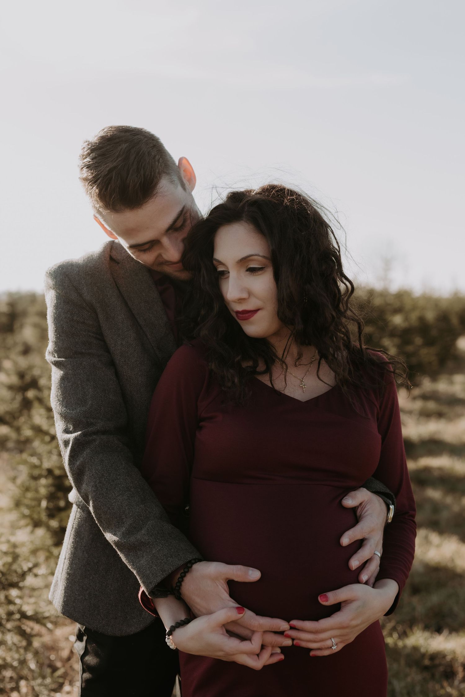 Man tenderly embraces pregnant woman in burgundy dress during outdoor maternity shoot.