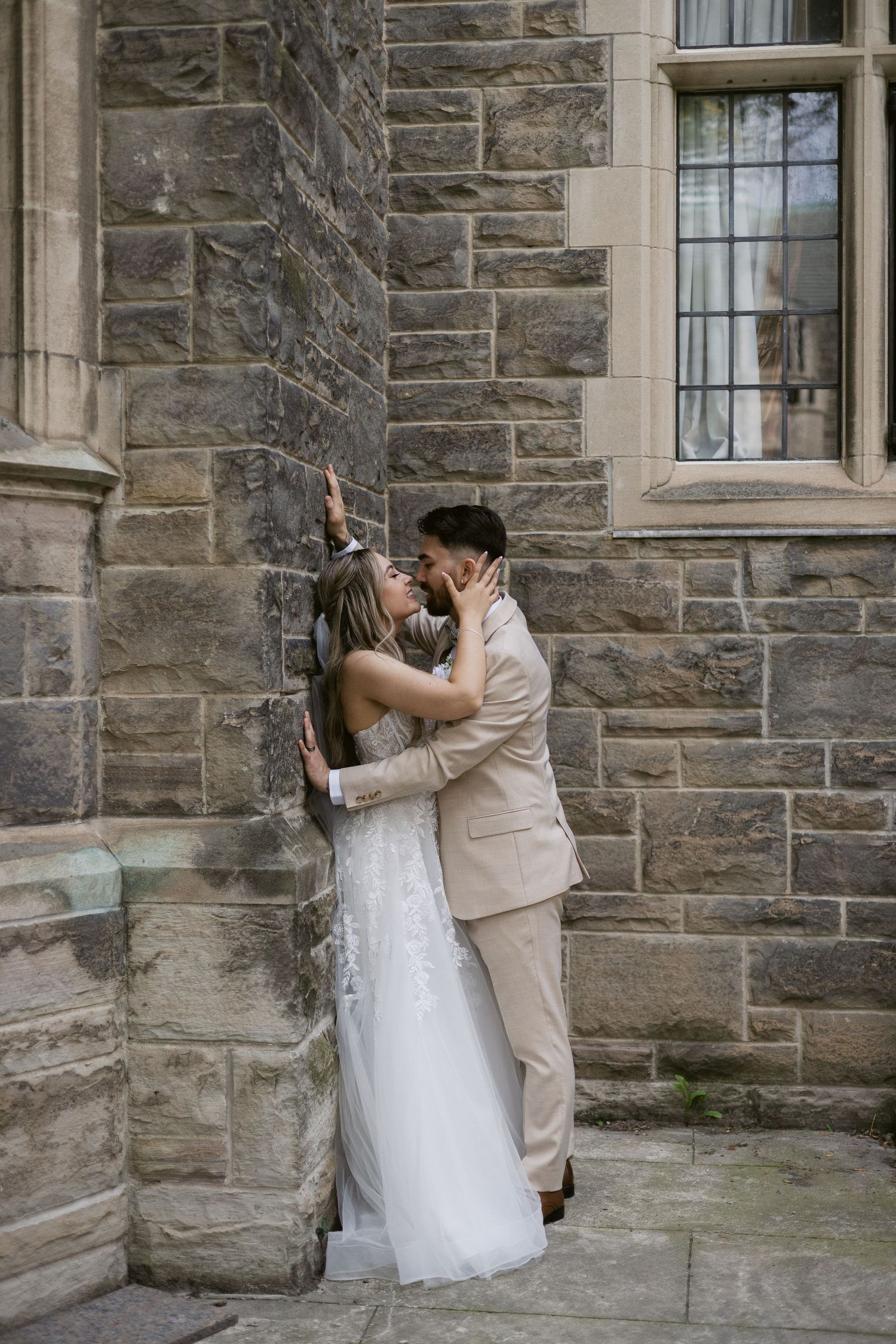 Bride and groom embrace and kiss against corner of historic stone building during wedding photo session.