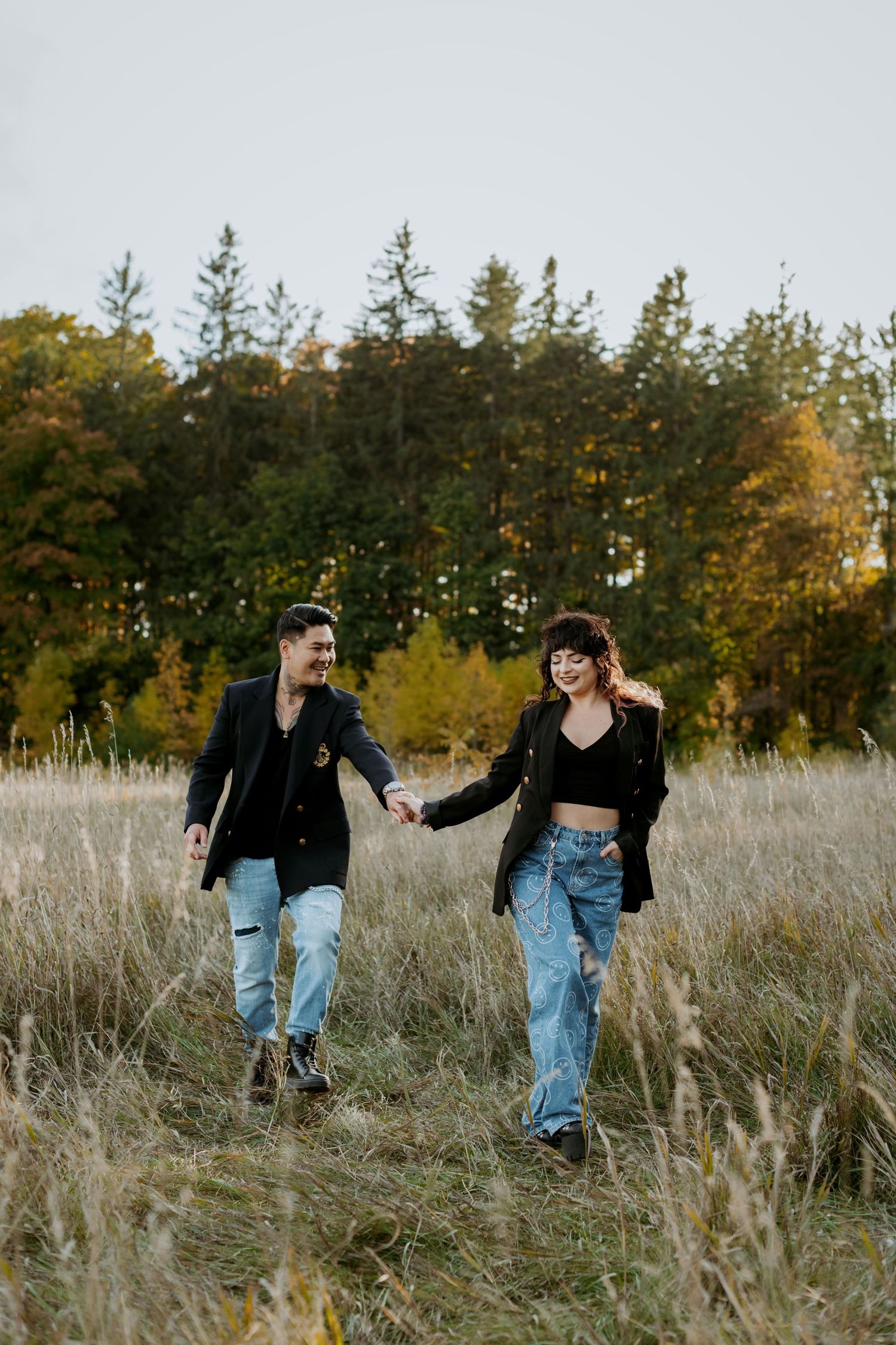 Happy couple in matching black blazers holding hands while strolling through golden grass field with autumn trees.